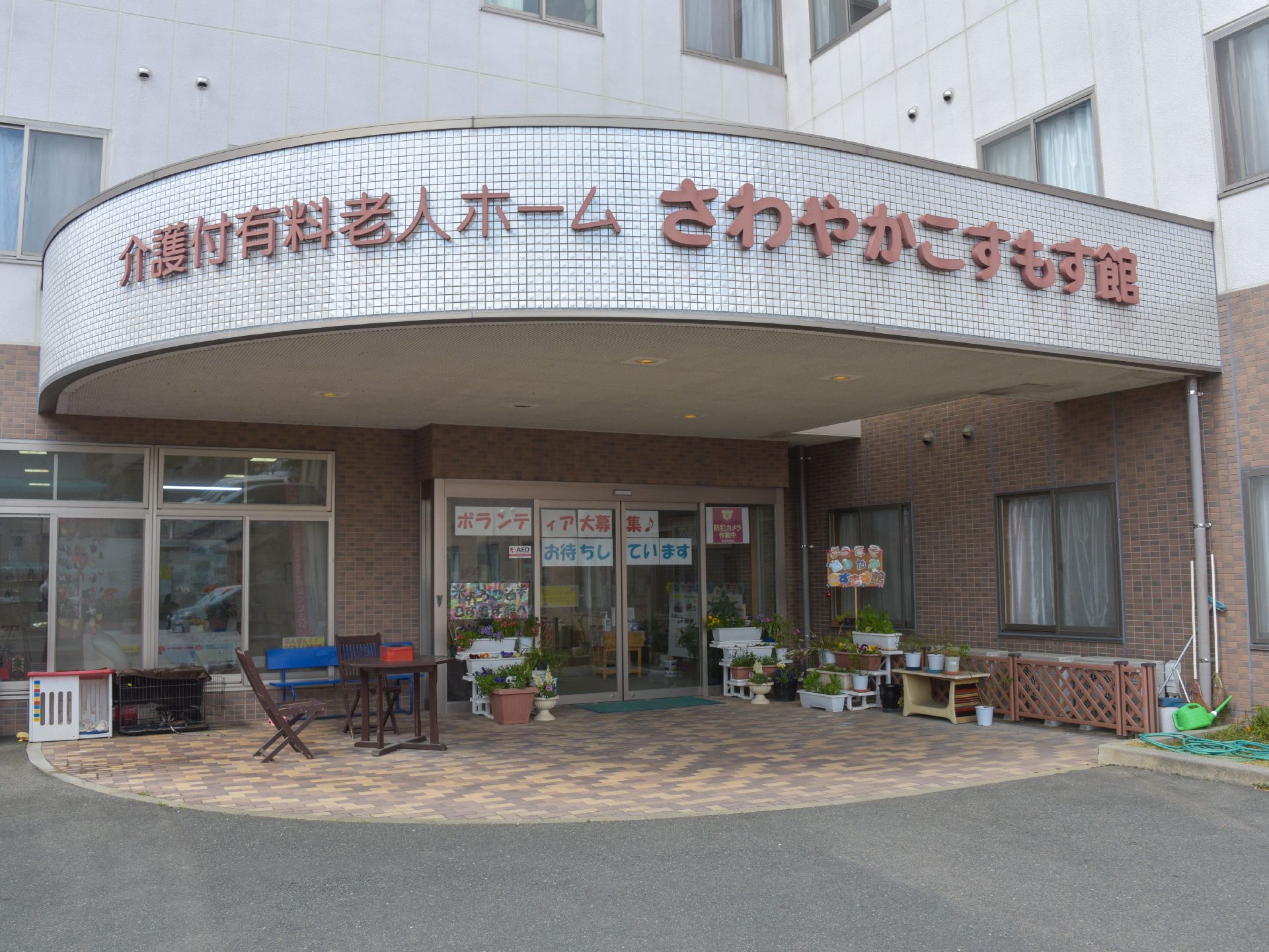 施設外観、玄関前の様子。施設名が掲げられている。自動ドアには「ボランティア大募集♪」の紙。多数の植木鉢や椅子とテーブルが設置。