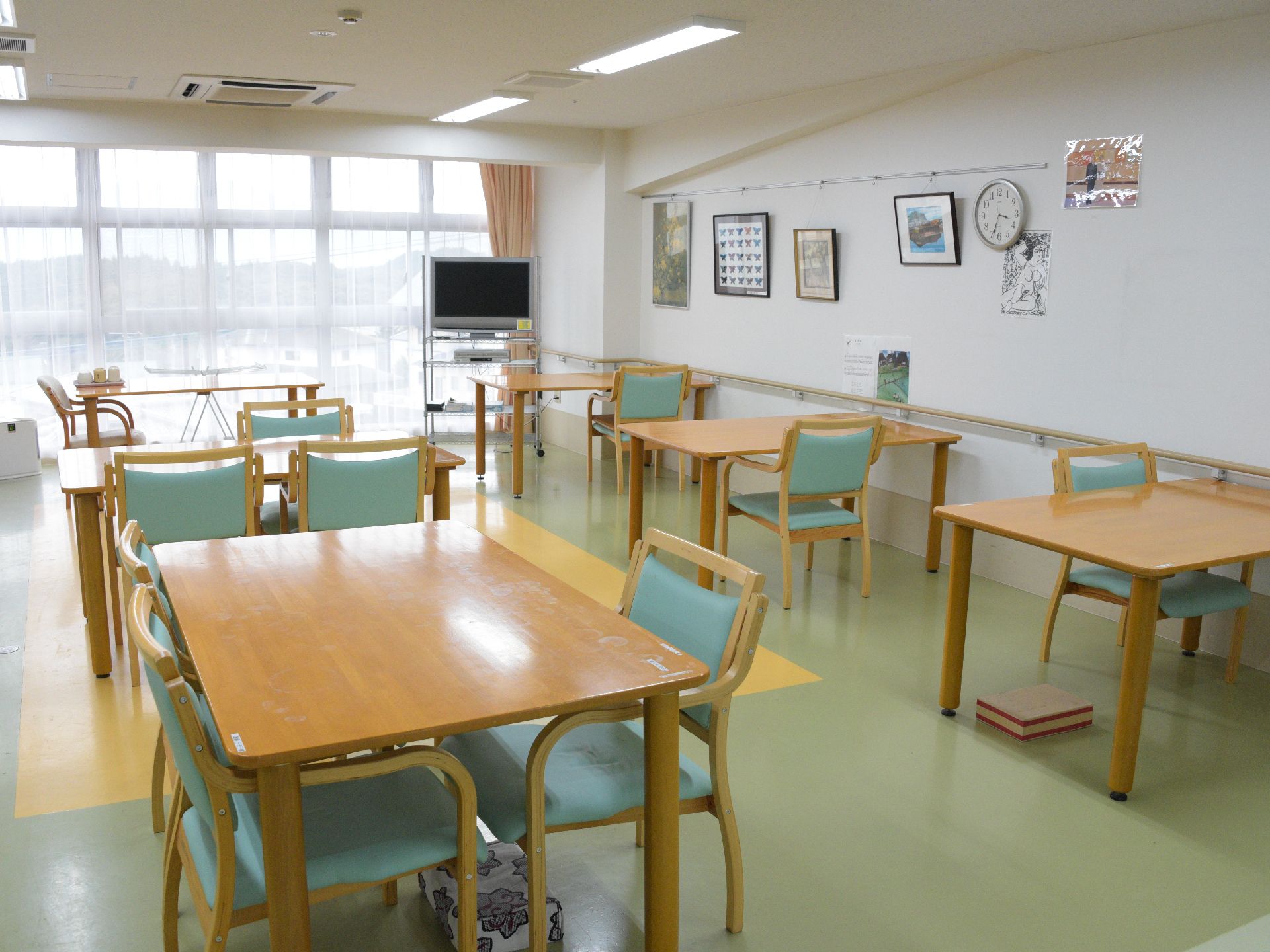 四角いカーキのテーブルと椅子が並んでいる。奥には白いカーテンがかかった窓が。床は黄色と緑。白い壁には手すりがついている。