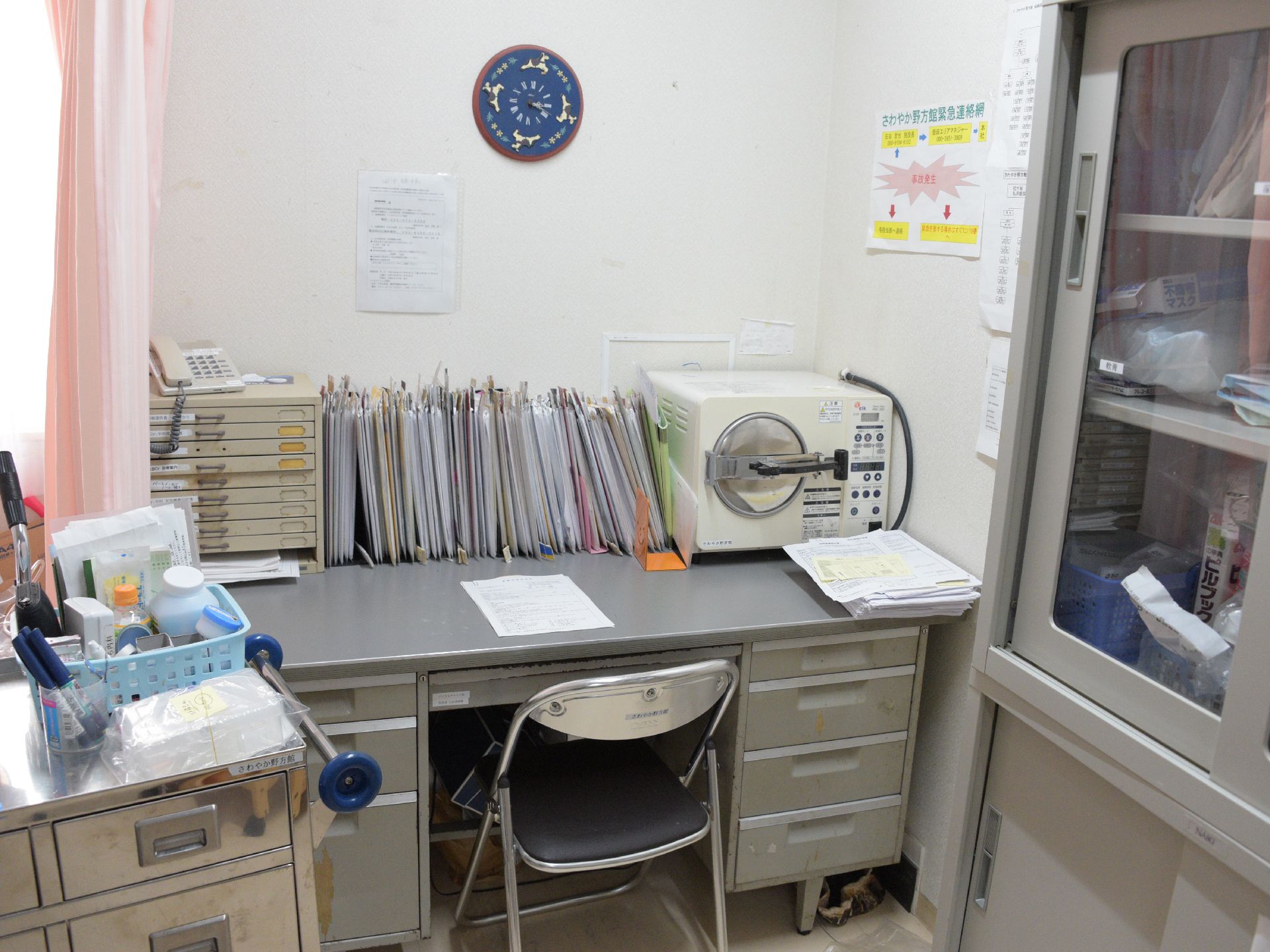 診察室の様子。白い壁には時計がかかっている。ディスクの上には電話と大量の資料、機材が。椅子はパイプ椅子。