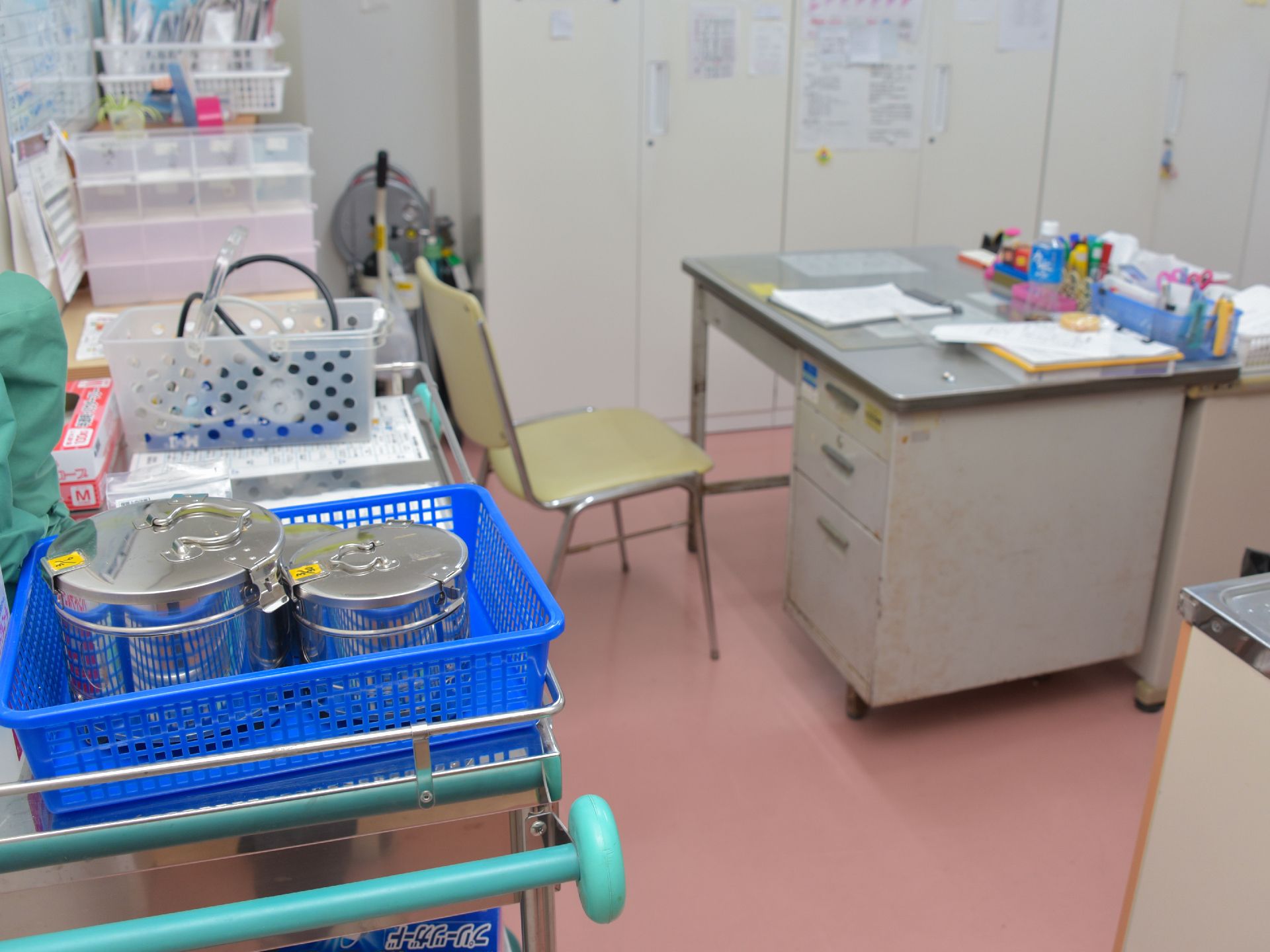 診察室の様子。手前には消毒ガーゼなどが入ってそうなステンレス容器が入ったカゴが。机と椅子。奥にはロッカー。
