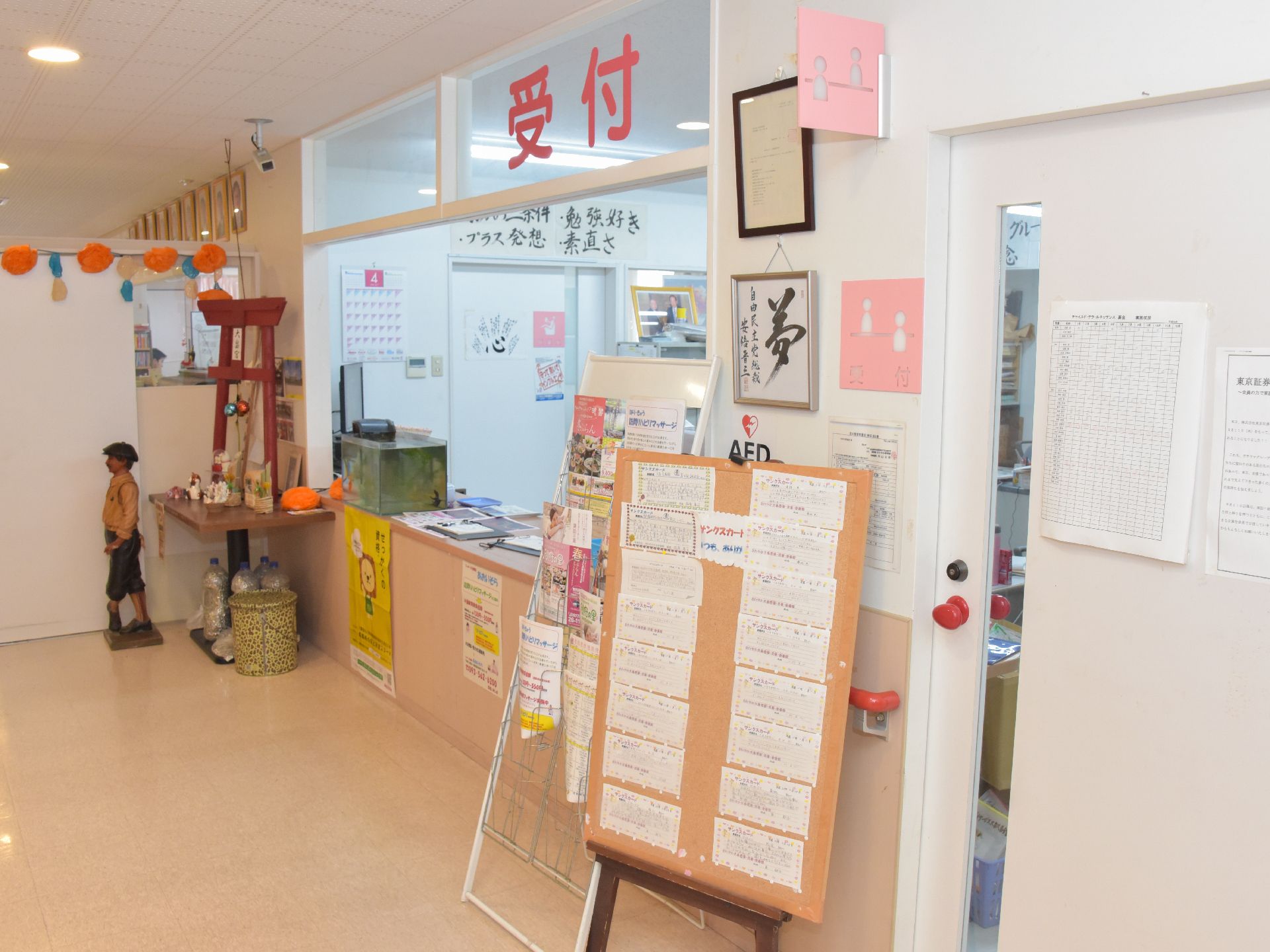 受付の様子。床は薄いベージュ。ボードや壁にはたくさんの掲示物が貼られている。奥には鳥居や人形が置かれている。