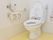 床はクリームイエロー。白いトイレ。壁には手すりとトイレットペーパーのホルダー、シャワートイレの操作ボタンと呼び出しボタン。
