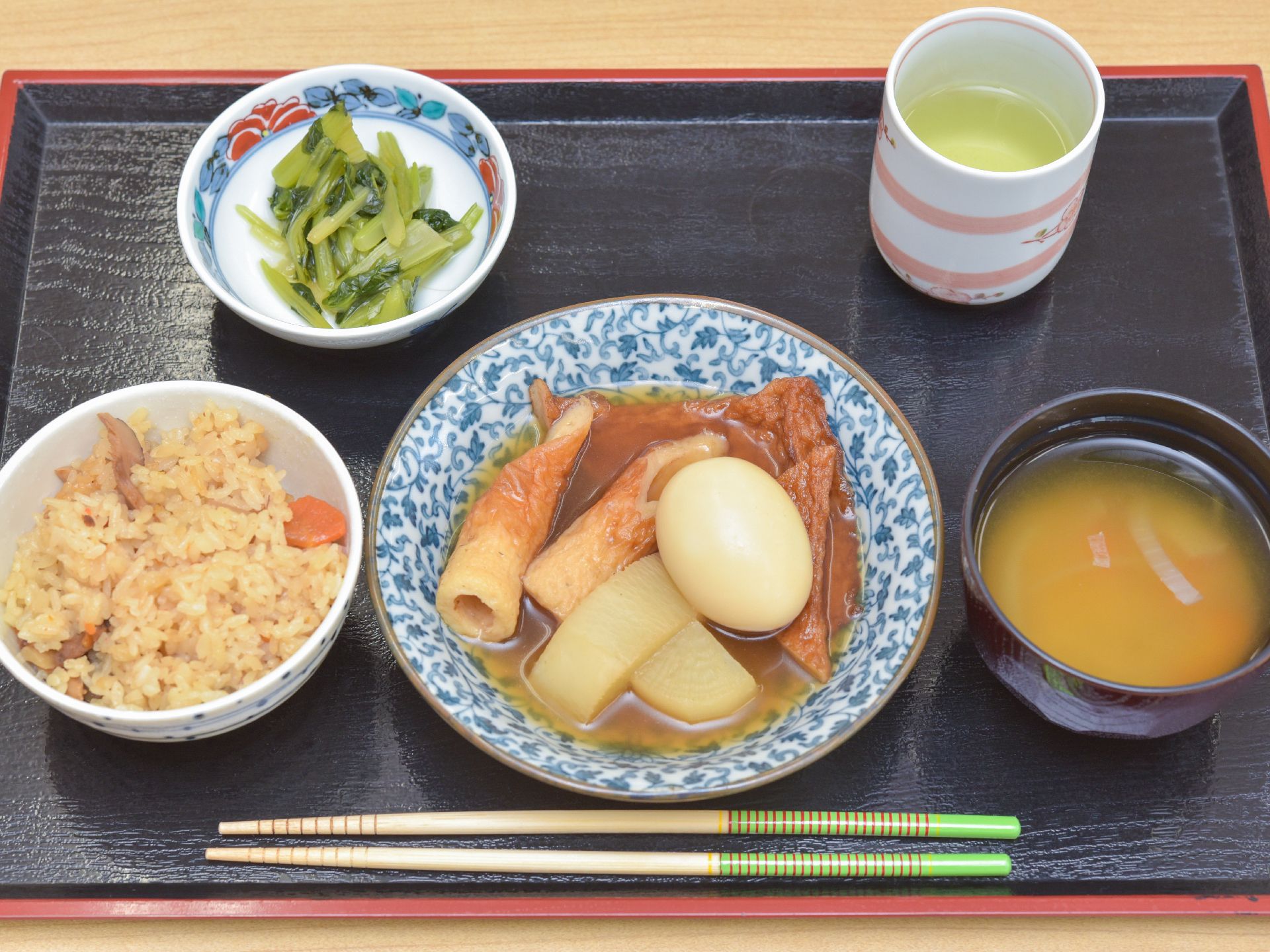 食事は、和食。黒いおぼんの上の器に、味噌汁・煮物・炊き込みご飯・お浸しが盛られている。飲み物にお茶がついている。