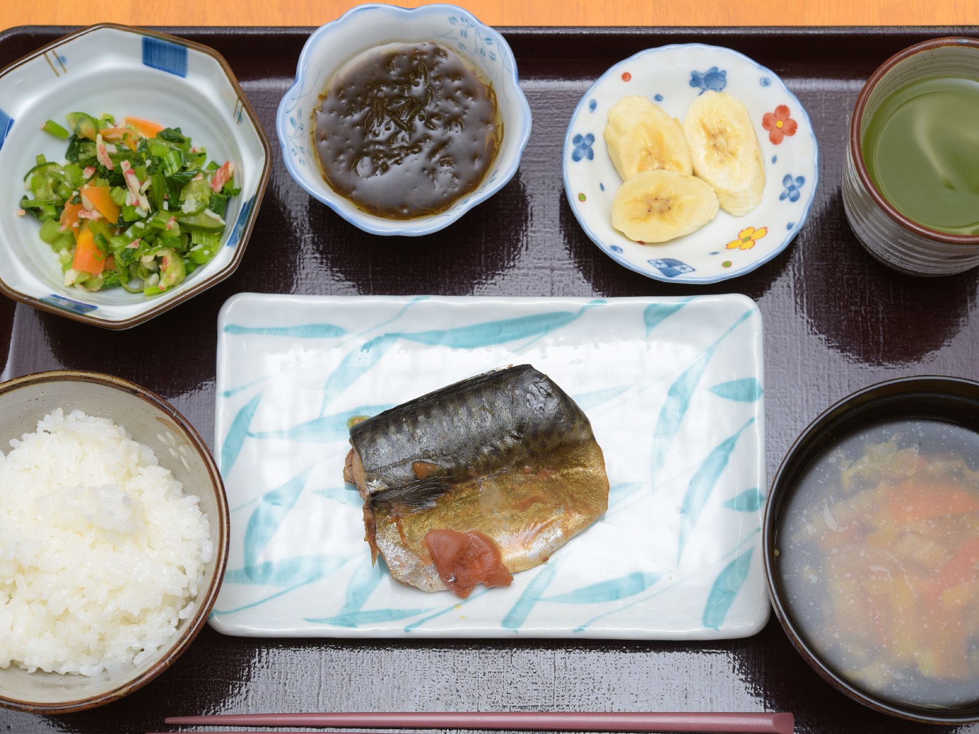 食事は和食。黒いおぼんの上には様々な器に、ご飯・味噌汁・お浸し・酢の物・お魚・バナナ・お茶が盛られている。