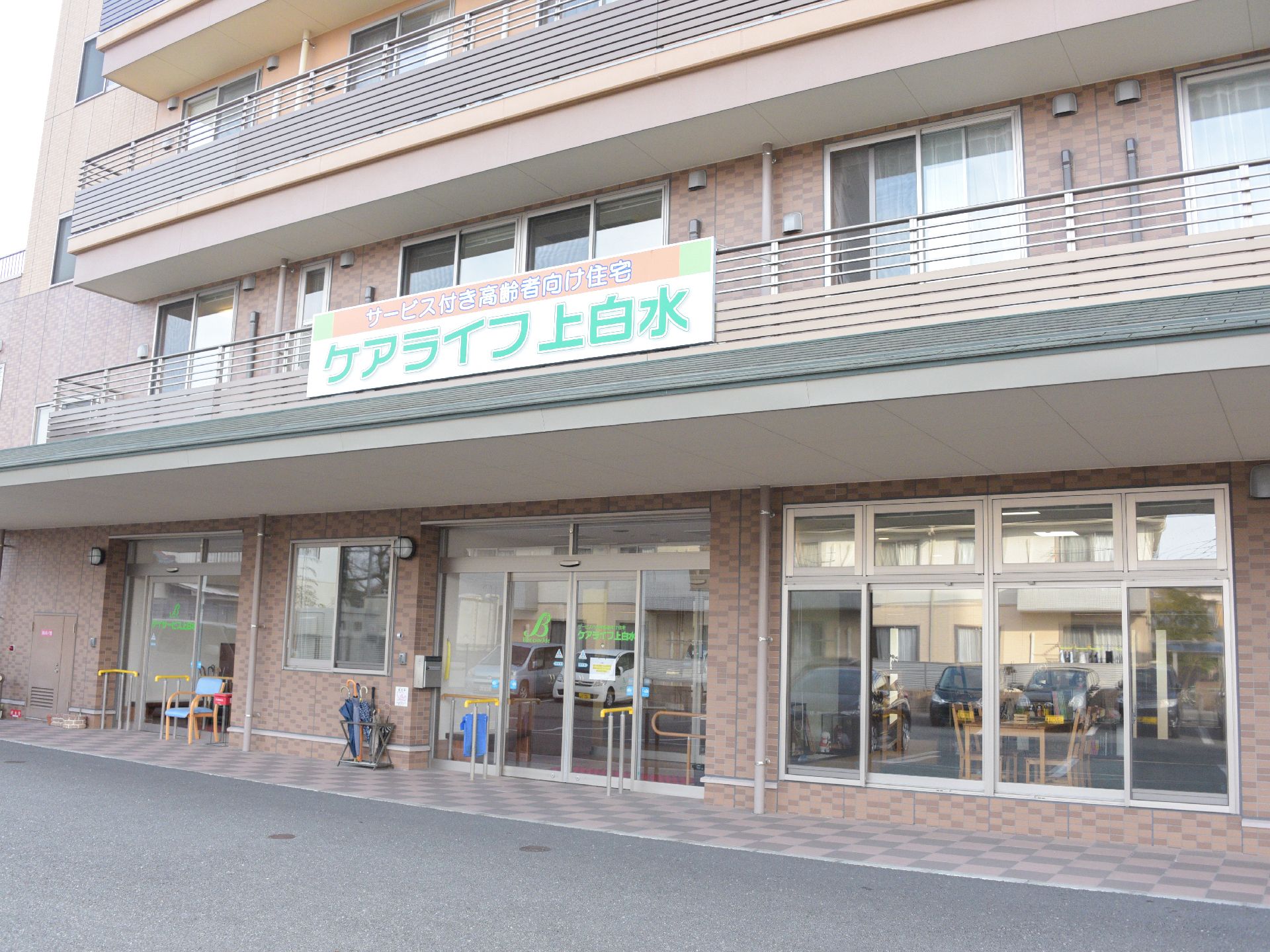 茶系のレンガ調の外壁の鉄骨造りの建物。玄関入口の上には施設の看板が掲げてあり、玄関ドアの前には手すりが設置してある。