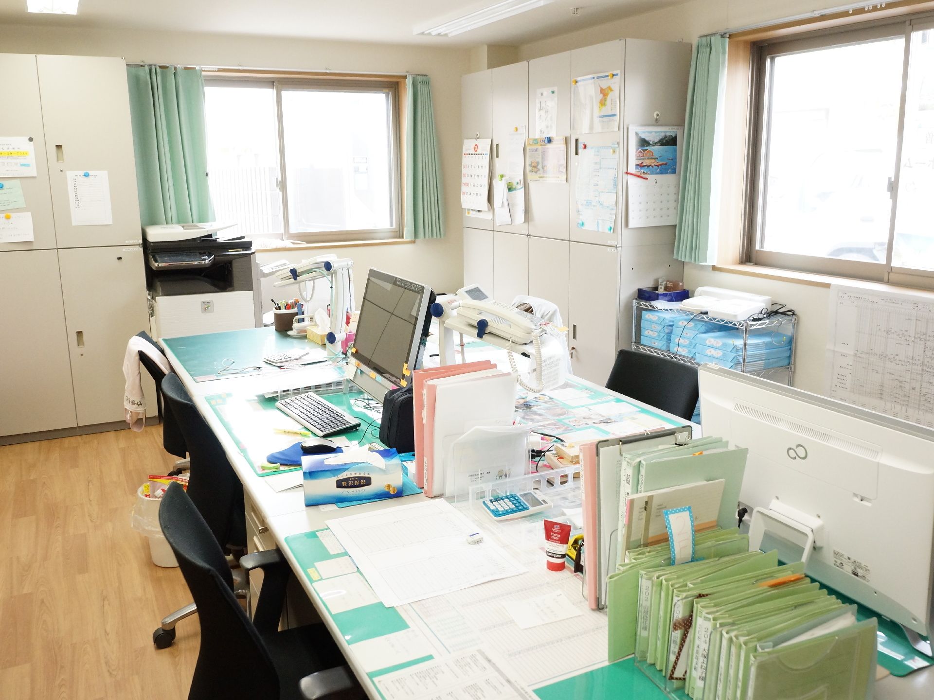 2つの窓から明るい光が差し込んでいる。机が向かい合わせで数台並んでおり机上にはパソコンや書類、ファイルなどが置かれている。