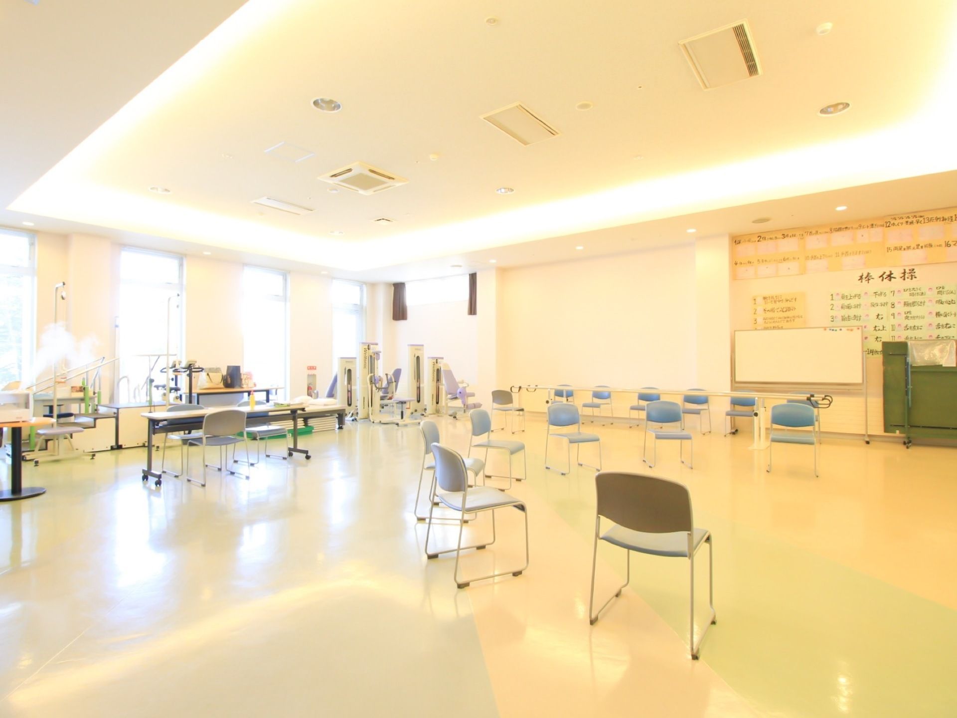 空調が整った開放感のある機能訓練室で、随所に器具が複数置かれているほか、壁には体操のポスターが貼られ、円形に椅子が並べられている。