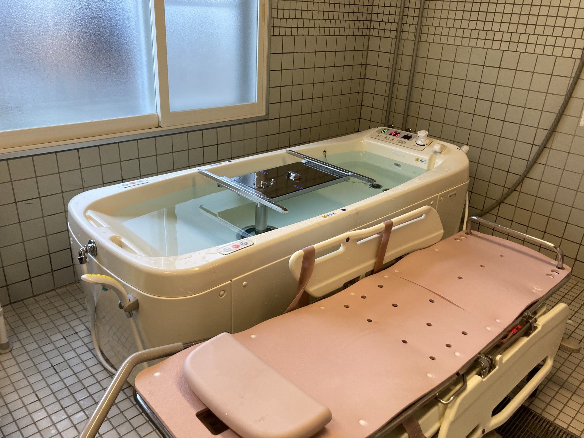 壁一面白色のタイル張りの浴室に、大きなピンク色の機械浴槽が設置されている。排水処理が施され、周りが濡れることがないように工夫されている。