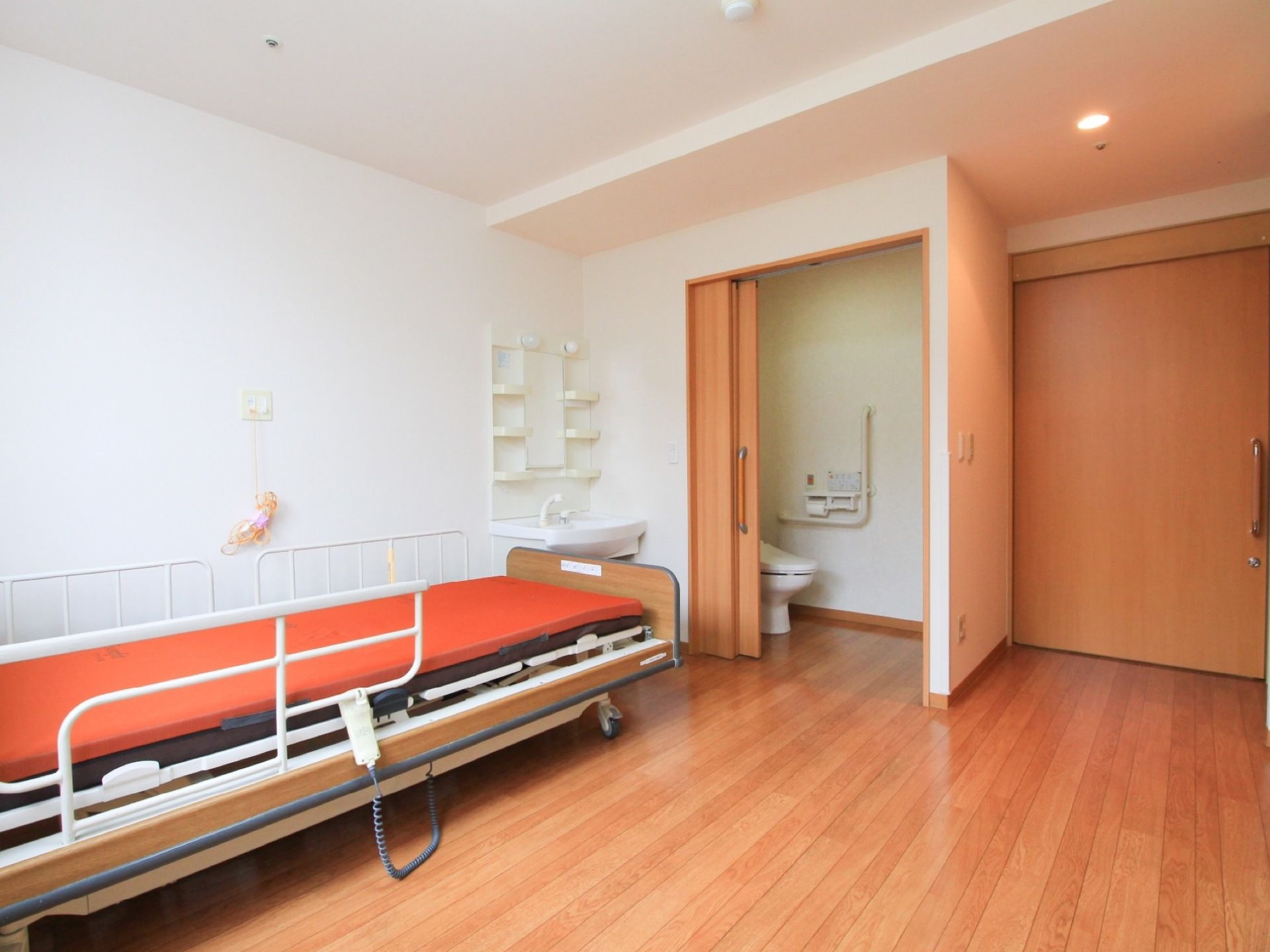 オレンジ色のマットレスが敷いてあるベッドが置いてある。トイレの扉が開かれ、白い洋式トイレが見えている。