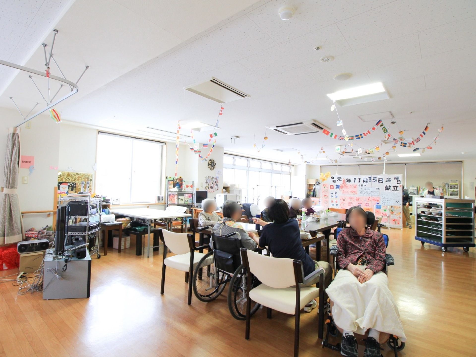 数名の入居者様が椅子や車椅子に腰を掛けている。キャスターが付いた配膳台が置いてあり、各棚には食器が入れられている。