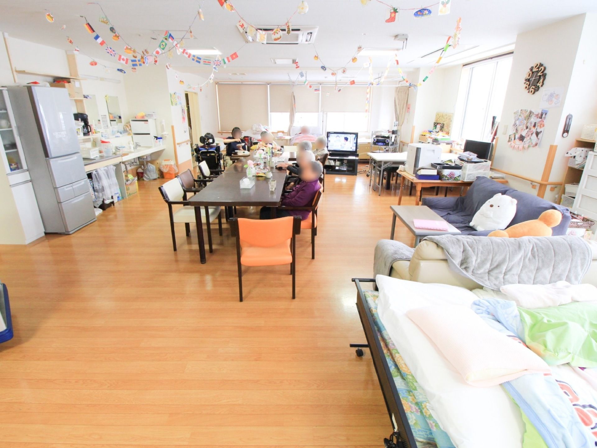 部屋の真ん中にダイニングテーブルが置いてあり、数名の入居者様が椅子に腰を掛けている。冷蔵庫や食器棚が壁際にある。