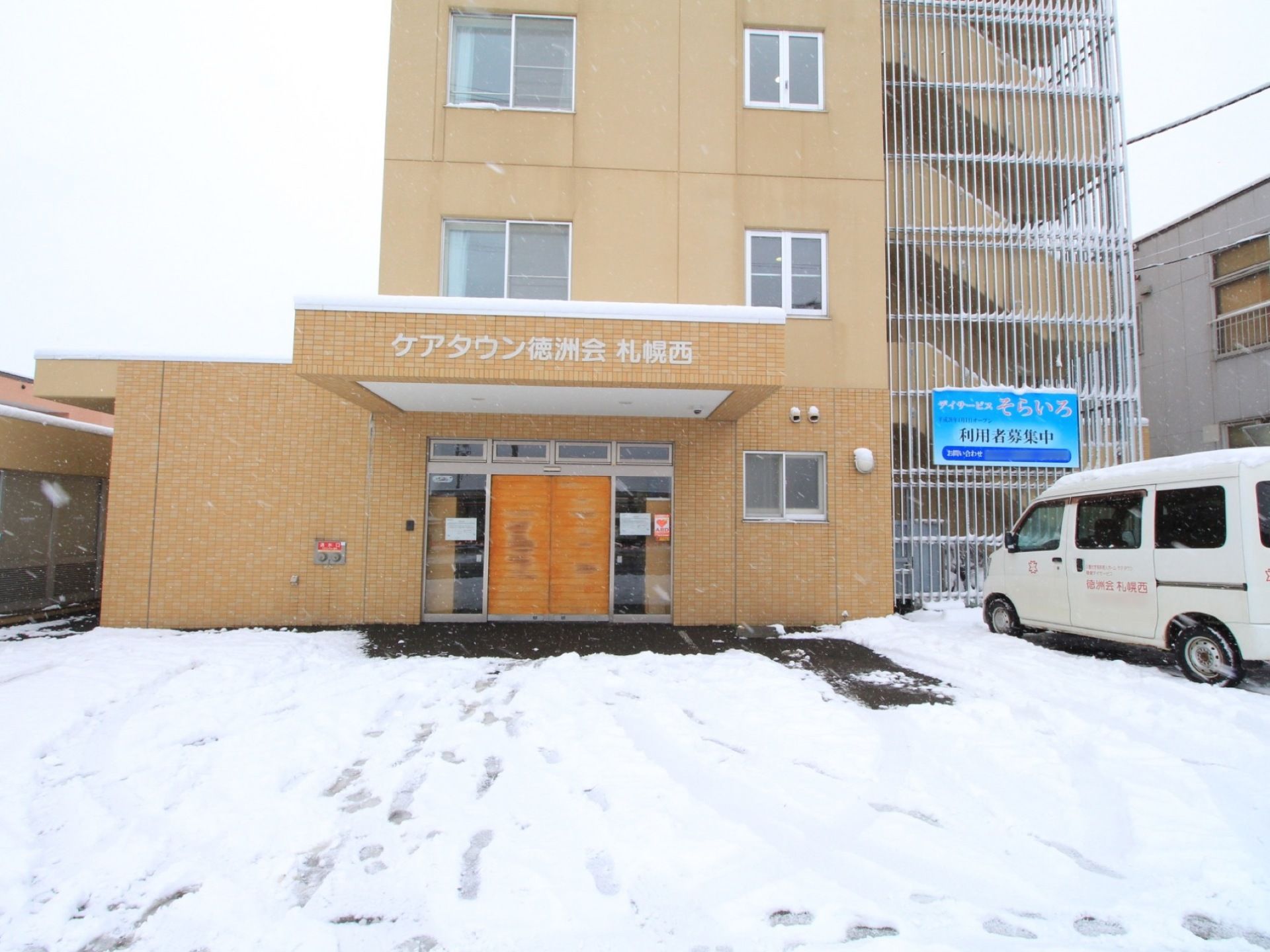 雪が積もった敷地内に白い車が停められている。屋根に施設の名称が表示され、木目の自動扉は閉められている。
