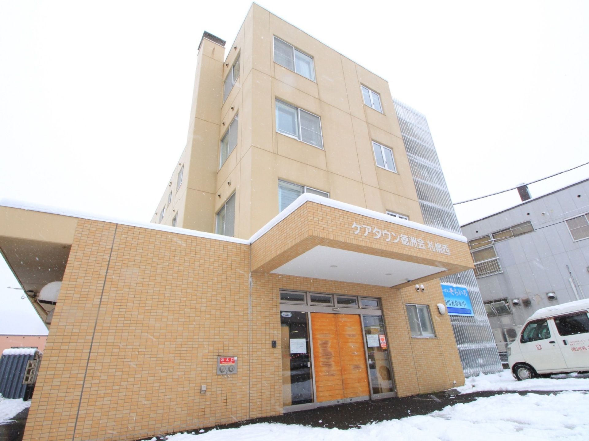 １階の外壁にレンガが貼られた４階建ての茶色い建物が建っている。玄関前は雪が積もり、白い車が停められている。