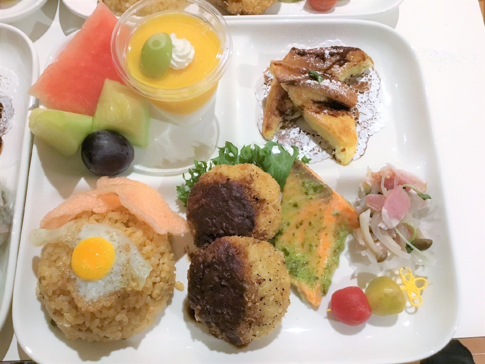 目玉焼きがのったおにぎりや一口ハンバーグ、カットしたスイカやメロンとプリンのデザートが付いたワンプレートご飯