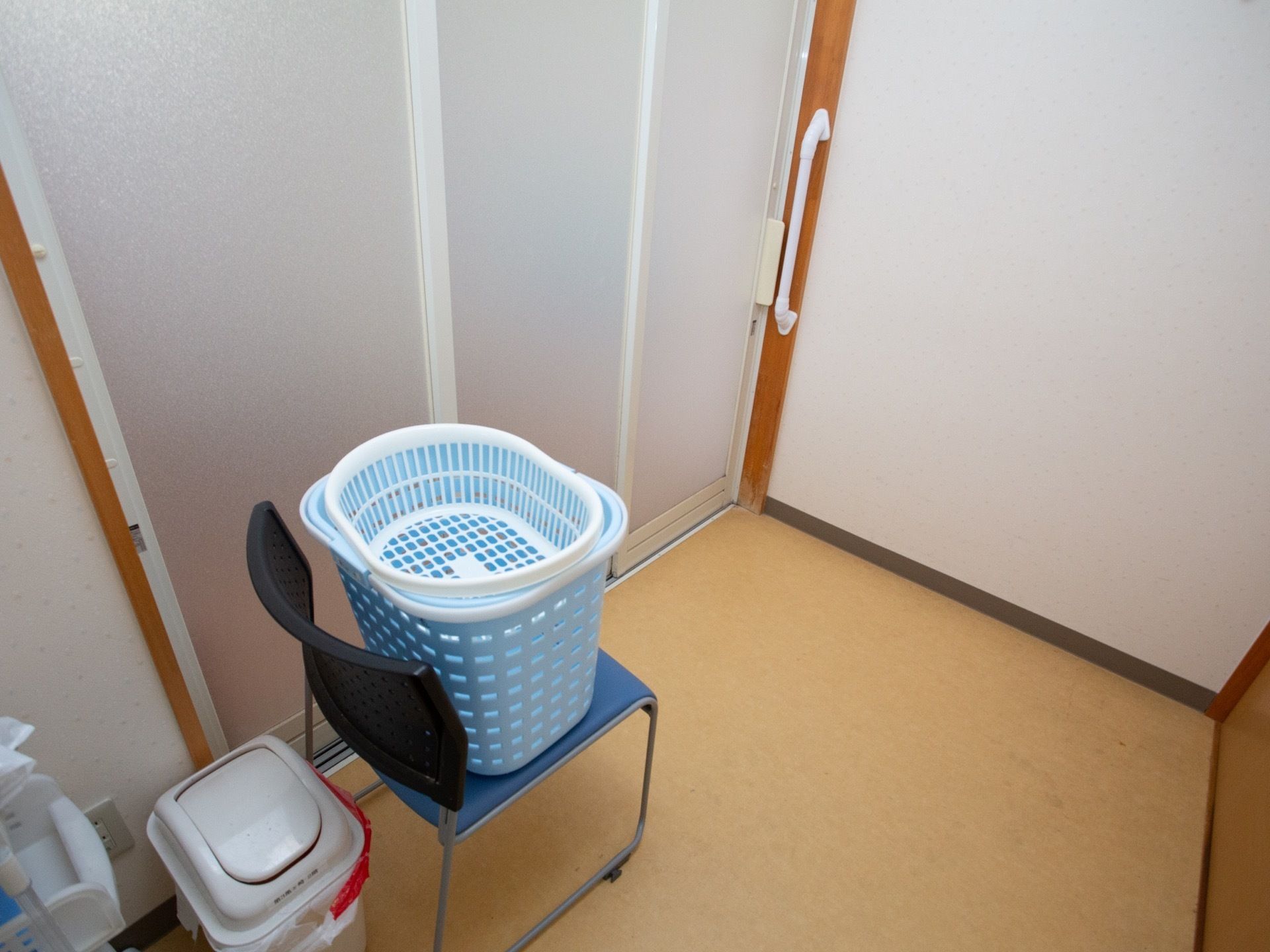 脱衣所のパイプ椅子の上に、青色と白色のかごが重ねて載せられている。その後ろには、ふたがついたごみ箱が置かれている。