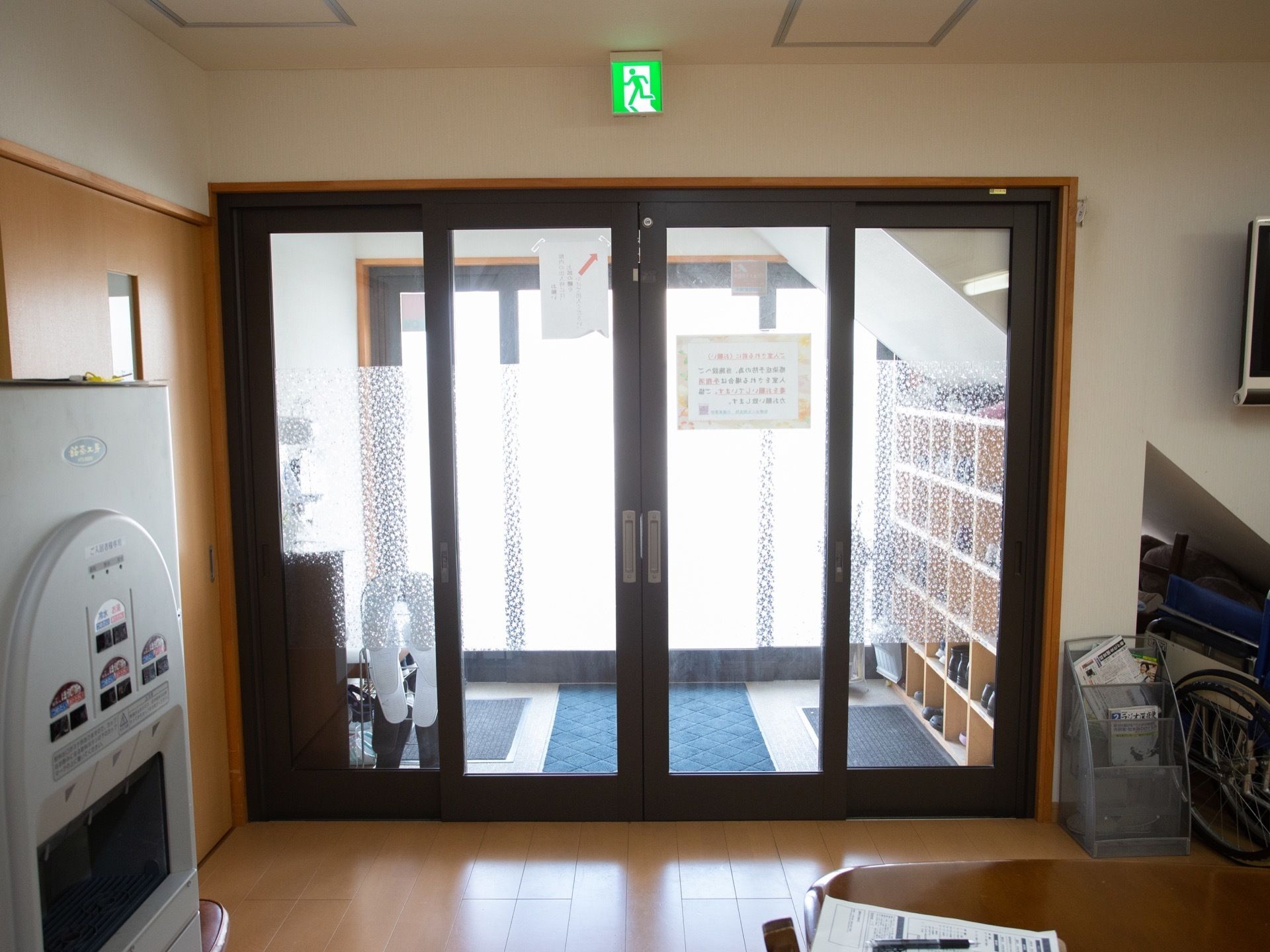 玄関を入った所に、水やお茶を提供するサーバーが設置されている。その前に新聞紙が置かれたテーブルがある。