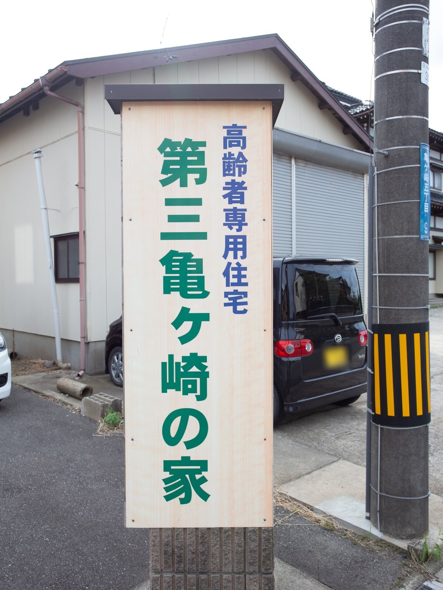 道路に立っている電柱のそばにあるブロックの上に、施設名称が大きな文字で書かれた縦長の看板が設置されている。