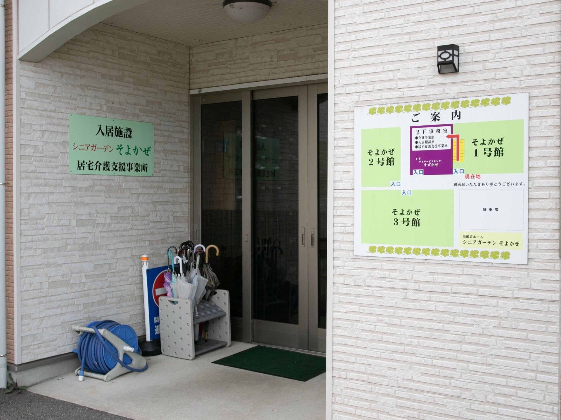 両開きの引き戸で、段差のない玄関がある。扉の横には傘立てやホースが置かれている。壁には施設全体の見取り図が掲示されている。