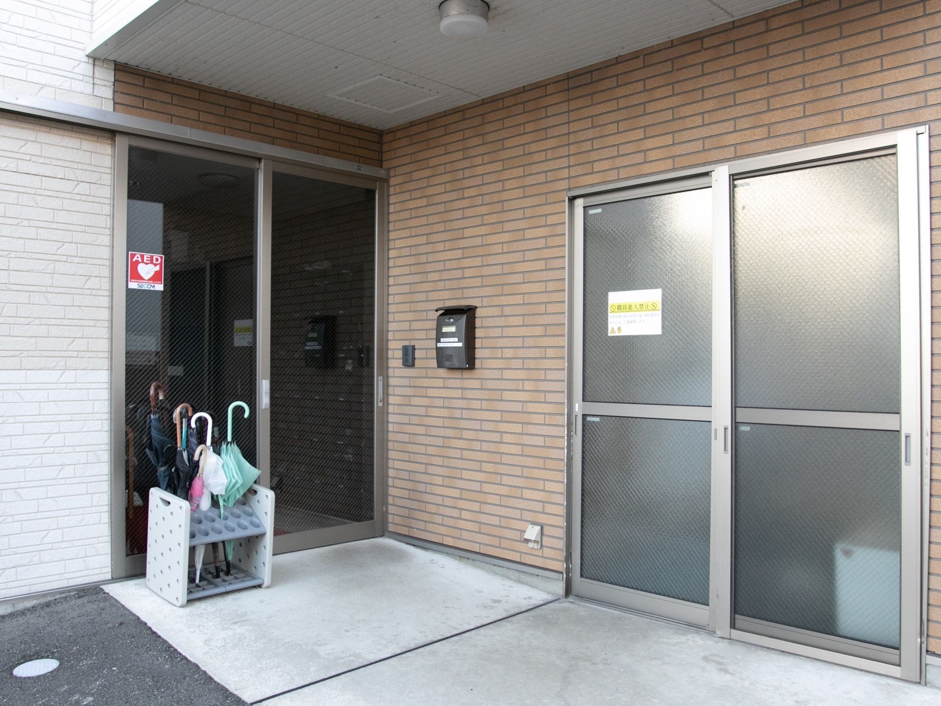 スライドドアの扉が２か所あり、一方にはＡＥＤのステッカーが貼られている。地面に置かれた傘立てに数本の傘が立てられている。