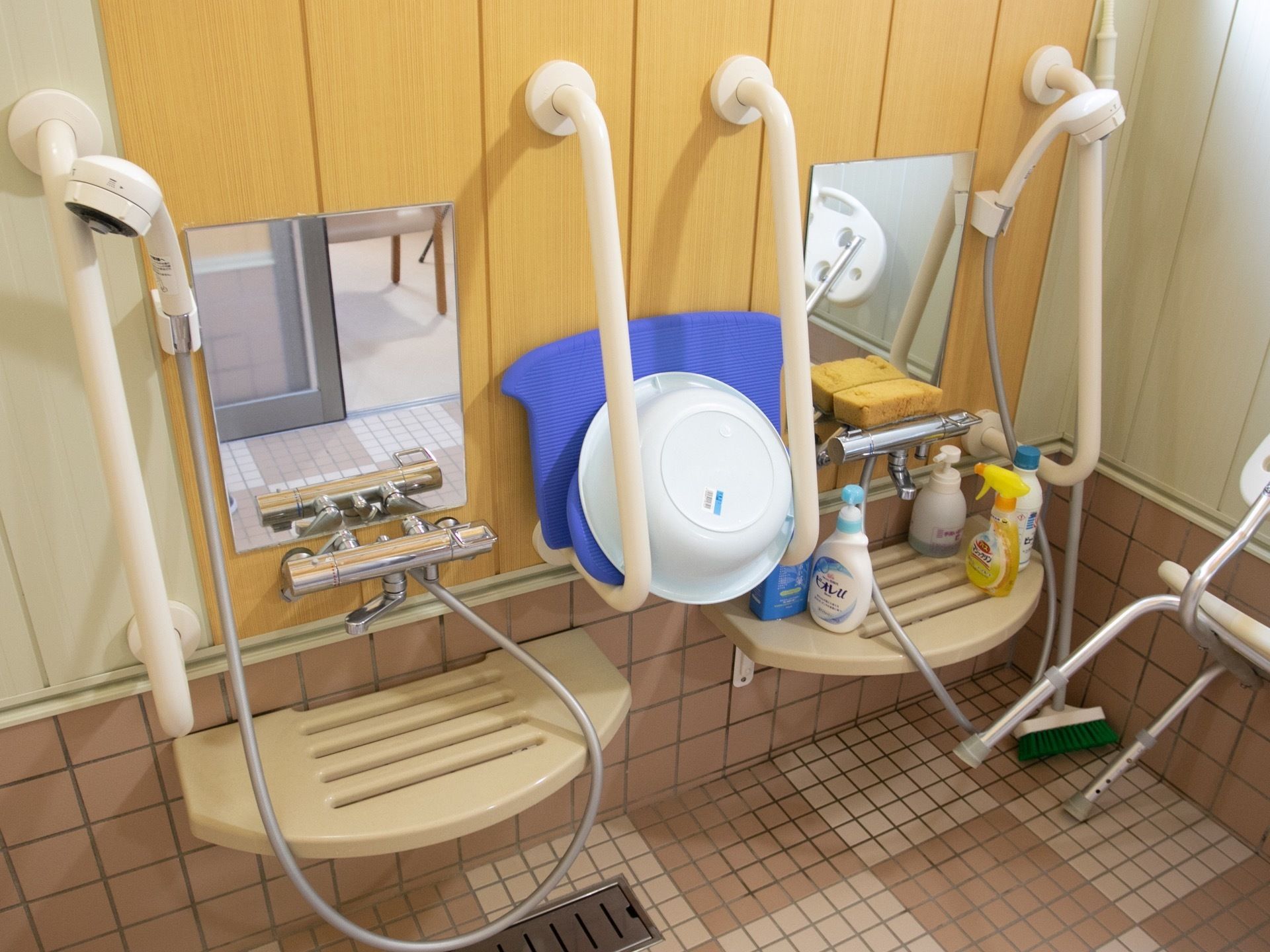 細かい目のタイルが敷かれた床に排水溝がある。壁にシャワーと鏡、手すりがある。白と緑色のデッキブラシが立てかけられている。