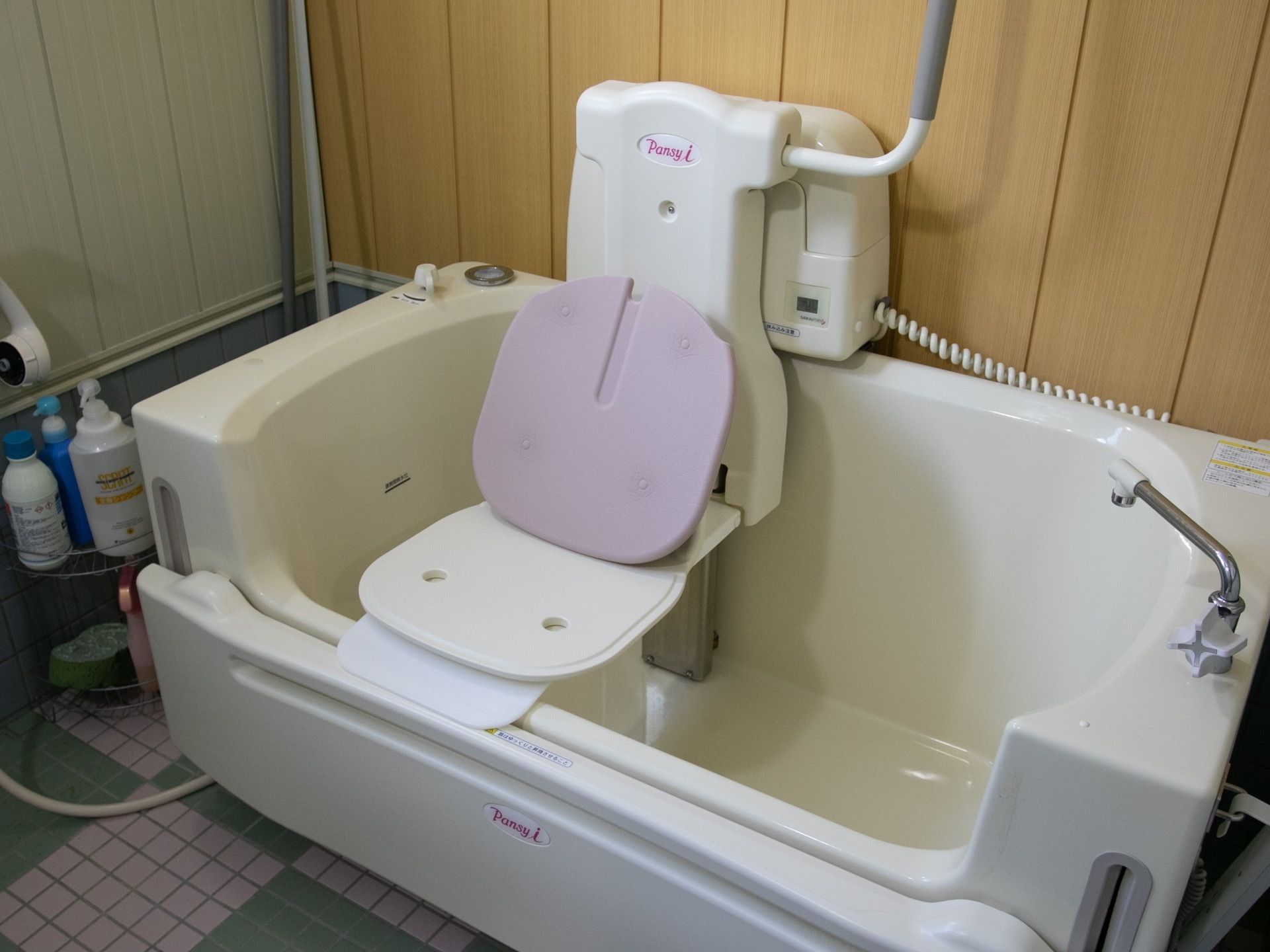 蛇口の付いた座位入浴が可能な介護浴槽がある。床は白色と緑色のタイルが敷かれ、浴槽の隣にシャンプーなどが乗った台が置かれている。