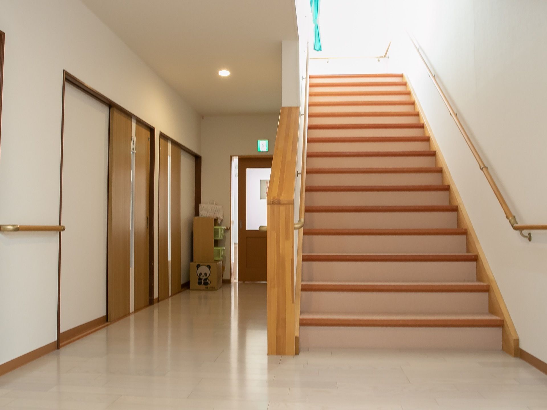 光が差し込む明るい階段がある。通路の奥にはパンダが表示された段ボールが床に置かれ、窓ガラスの付いた扉が少し開いている。
