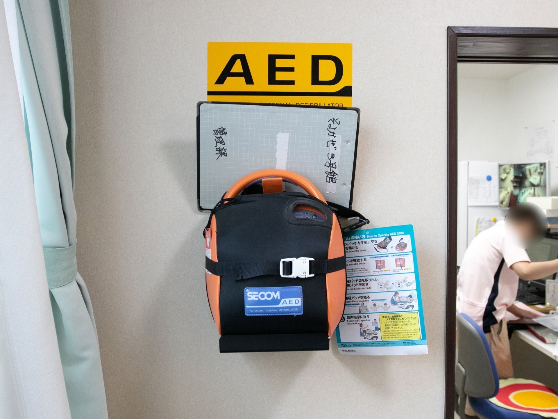 水色のカーテンとレースカーテンが掛かっている横の壁に、AEDの機械が掛けられている。扉が開いた部屋にスタッフがいる。