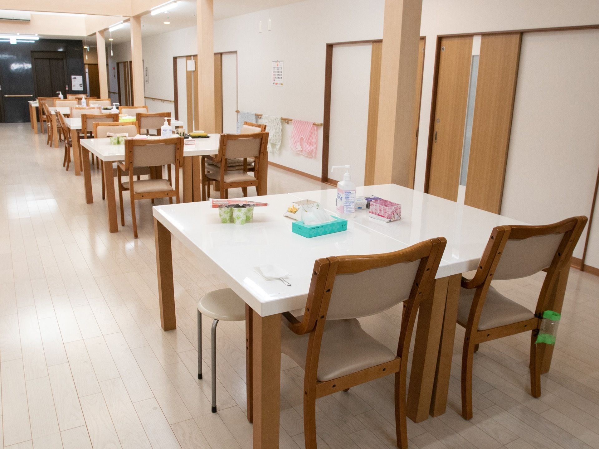 薄いベージュ色のフローリングの床の部屋に、たくさんのダイニングテーブルが並べられている。柱の奥には壁があり、手すりがある。