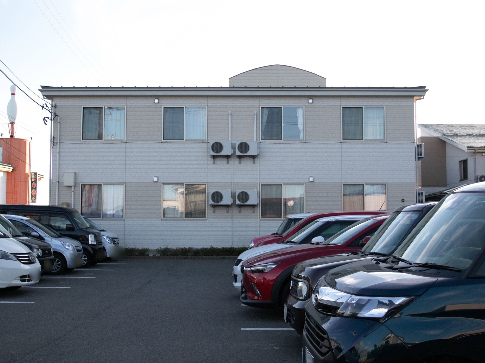 白い２階建ての建物があり、アスファルトで整備された駐車場にたくさんの車が停まっている。ボーリングのピンが乗った建物が建っている。