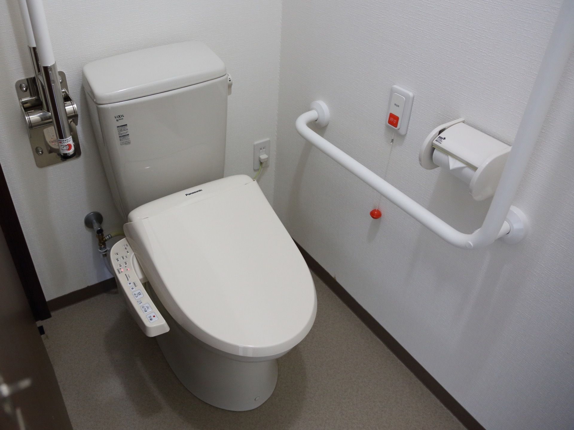 温水洗浄便座のついたトイレである。壁には手すりやナースコールが付いている。便器脇には跳ね上げ式の手すりがある。