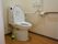 トイレ個室内である。温水洗浄便座付きトイレで、左手の壁に操作ボタンがある。また、壁に手すりが付いている。