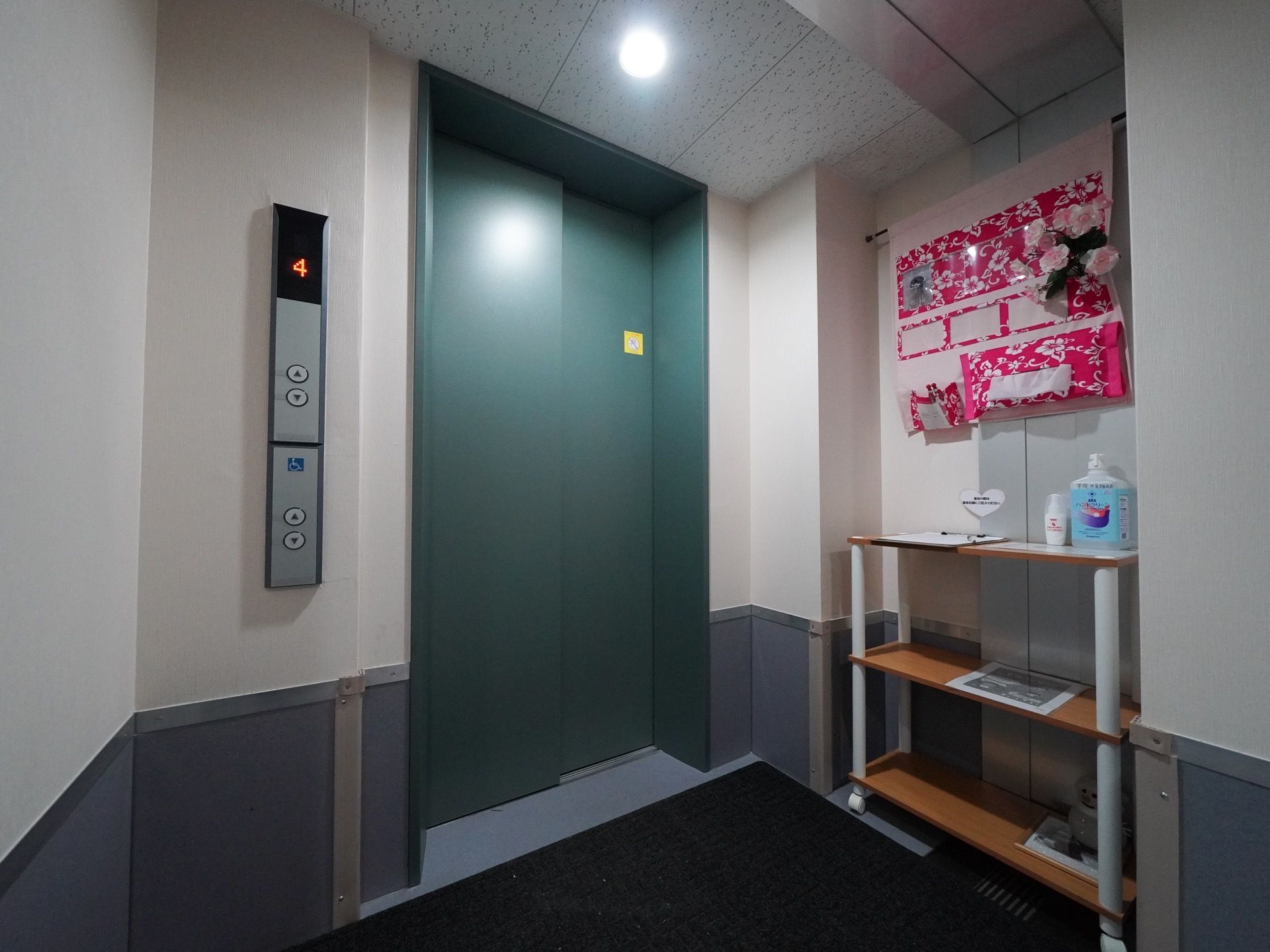 緑の扉のエレベーターである。扉の横に棚が置かれ消毒用スプレーが置かれている。その上に花柄のポケットが下げられている。