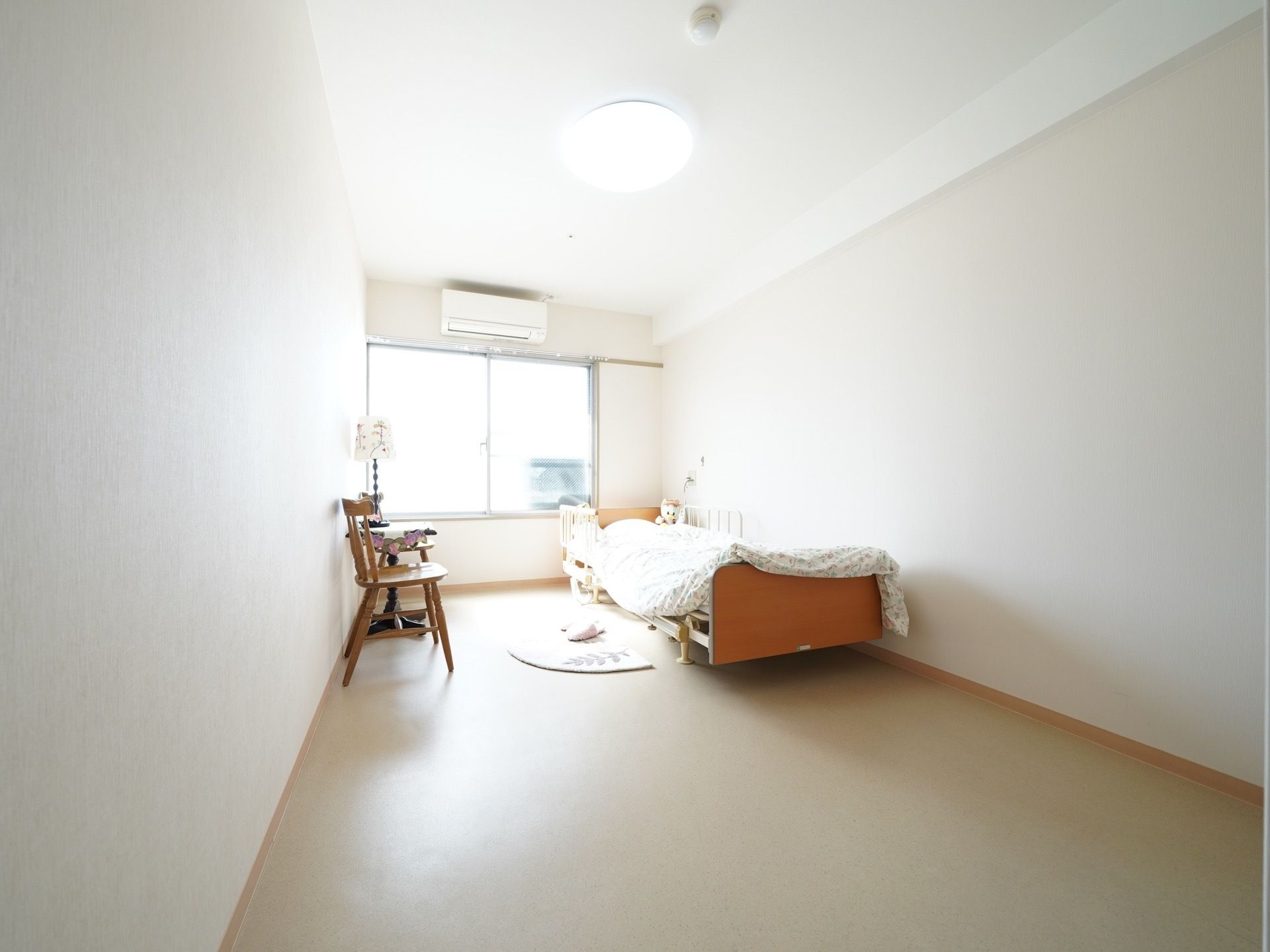 居室の奥に大きな窓があり、その上にエアコンが取りつけられている。ベッドの前にラグが敷かれ、テーブルセットが横にある。
