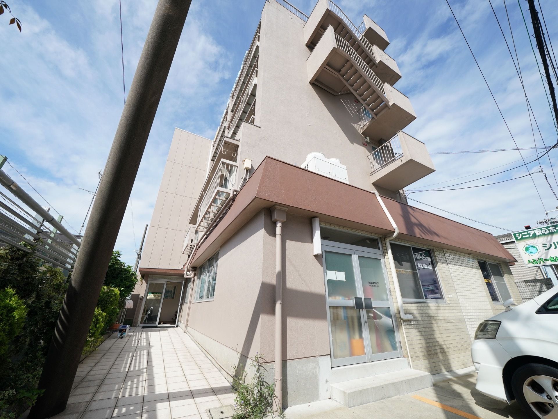 ベージュと茶色の建物で施設の前に車が１台停まっている。通りに向けて施設名の書かれた看板が立っている。