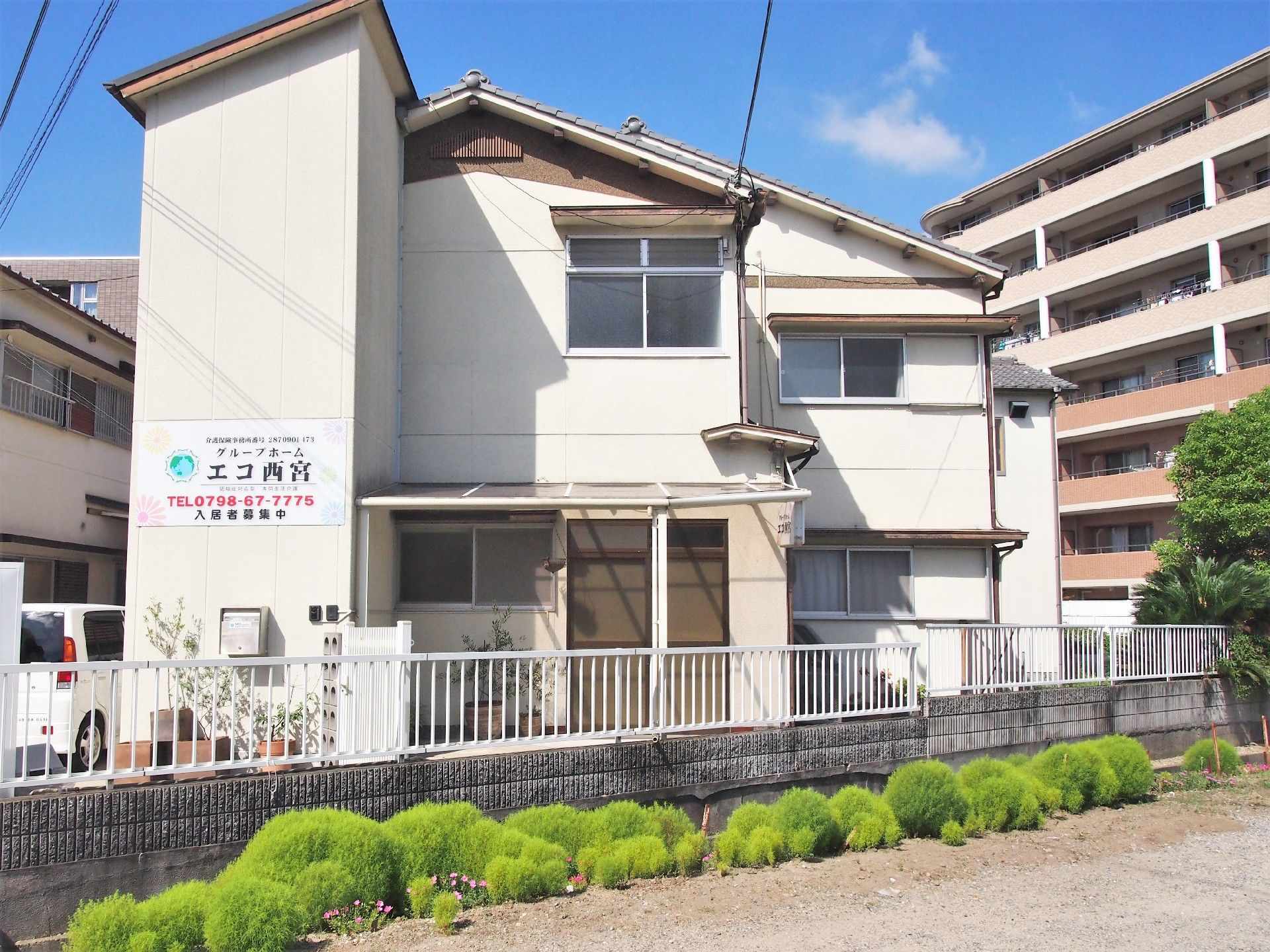白い外壁の２階建ての瓦屋根の建物である。玄関の前には門があり、その両脇に駐車、駐輪スペースが設けられている。