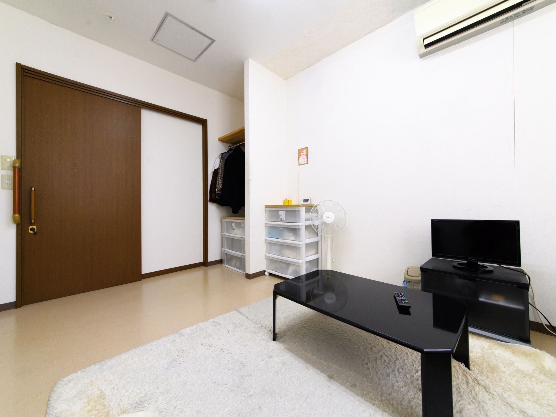 居室の様子。部屋にはテーブルや衣装ダンスなどが置かれている。ジュータンが敷かれている。テレビ、エアコン、扇風機などの家電もある。