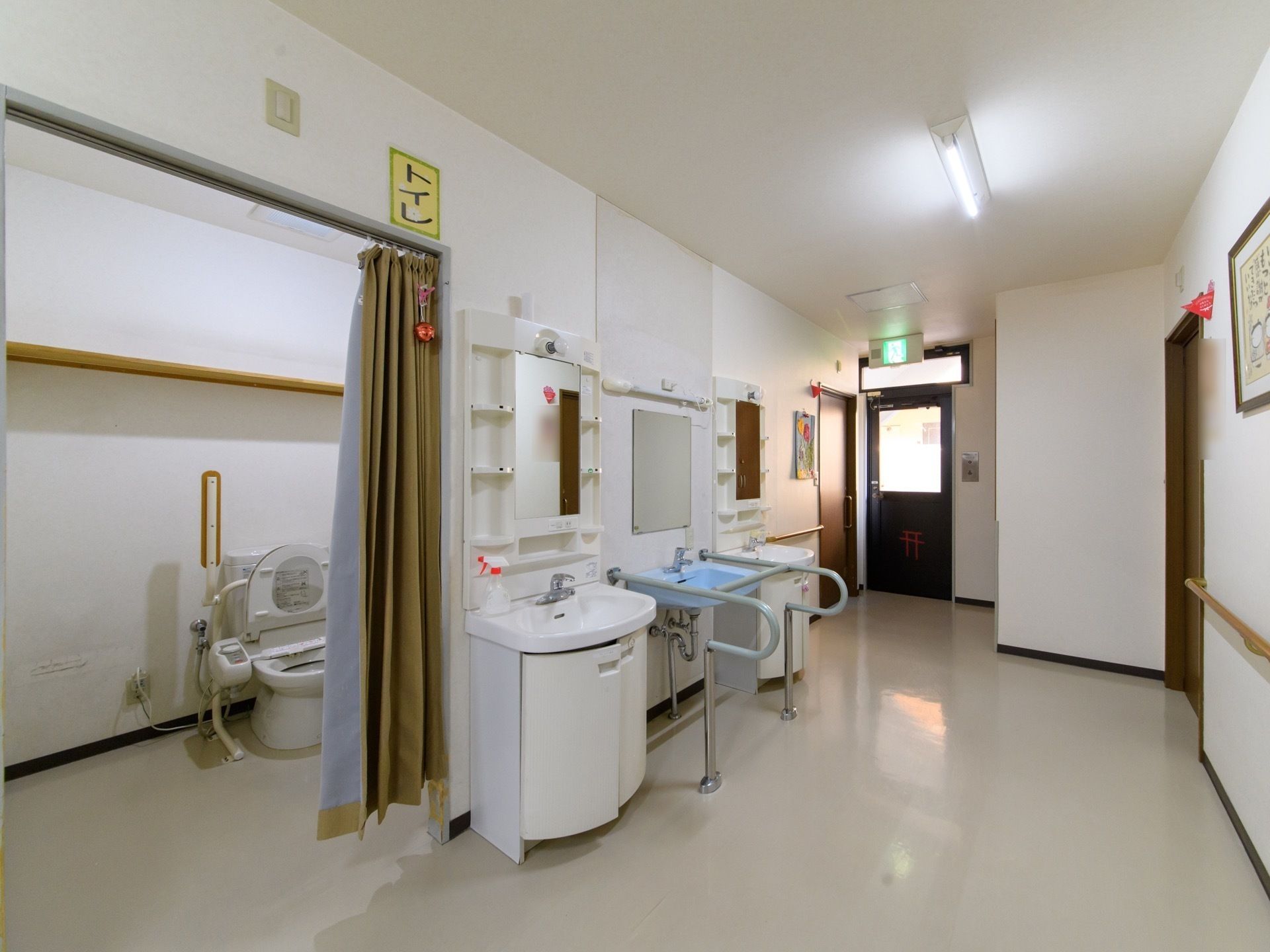 フラットな段差のない床面に洗面台が3台見られる。真ん中の洗面台は、コの字型の手すりが付いている。奥にトイレが見える。