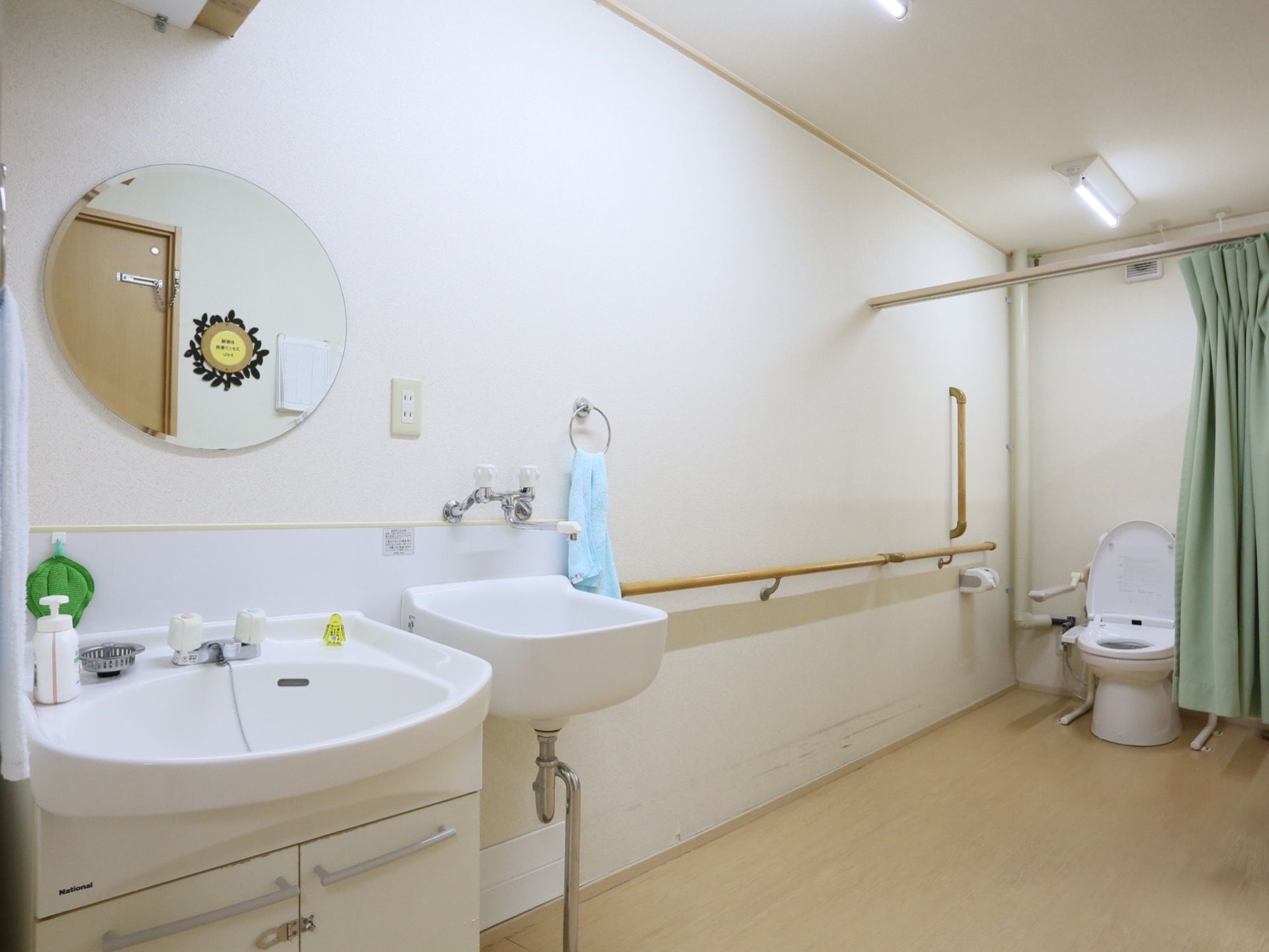 明るい色合いの居室である。丸型の鏡が備えつけられた洗面台がある。トイレの出入口にはグリーンのカーテンが設置されている。