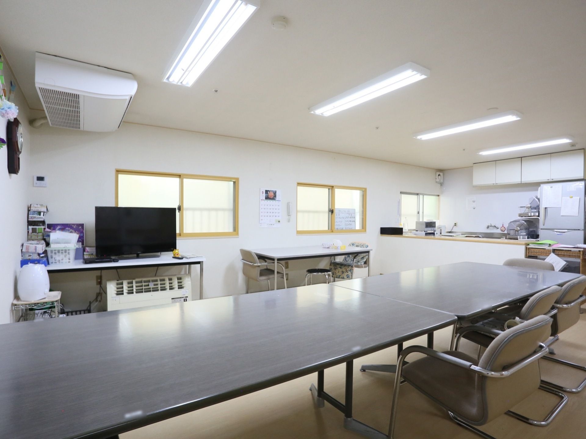 落ち着いた色あいの長机に椅子がついている。対面式カウンターがありキッチンが見える。広い部屋にはテレビも設けられている。