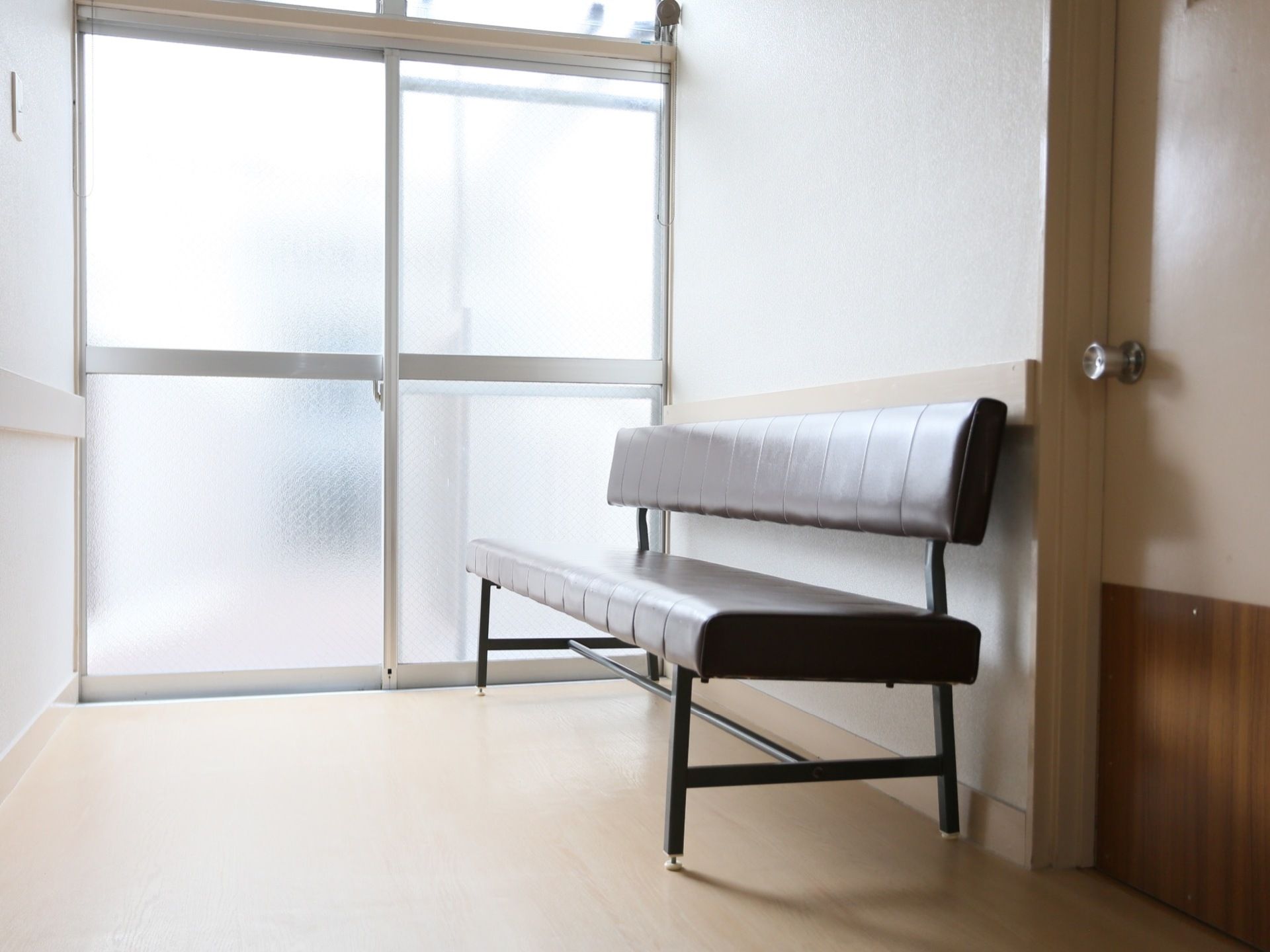 広々とした掃き出し窓が備えつけられており、外の日差しが通路に降り注いでいる。壁側には、背もたれつきのソファが置かれている。