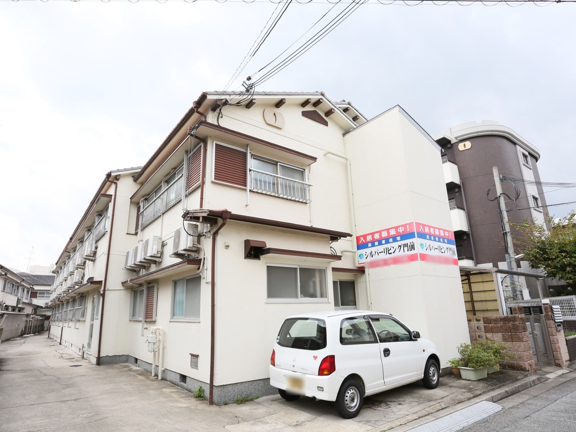 玄関前には一台の白い車が停まっており、木造二階の当施設が向かい側にも並んでいる。周囲は静かな住宅街で近くにコンビニエンスストアがある。