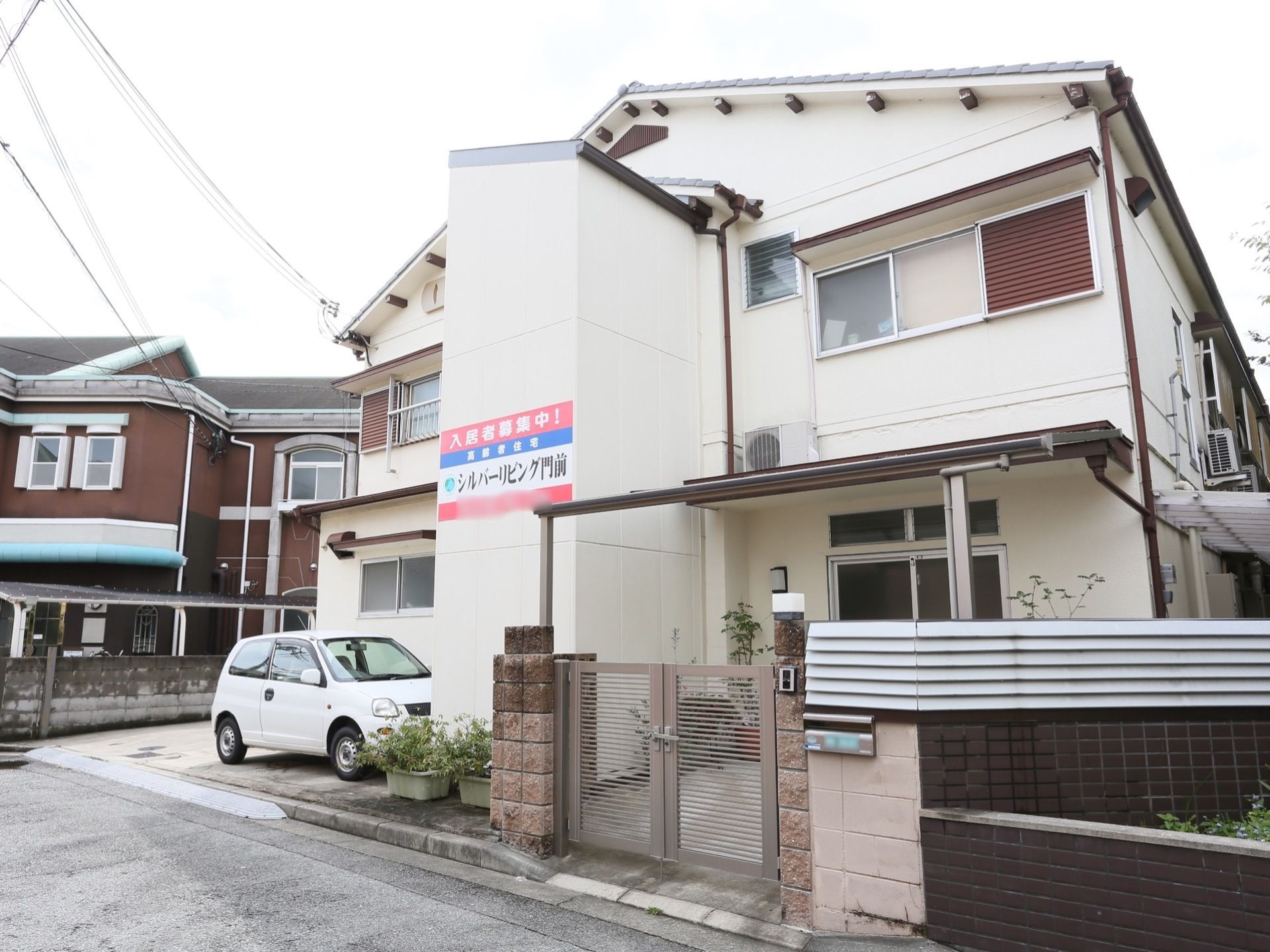 施設は道路に近い場所に建てられている。建物前に車一台分の駐車スペースがある。玄関前には、低めの塀と門扉がある。