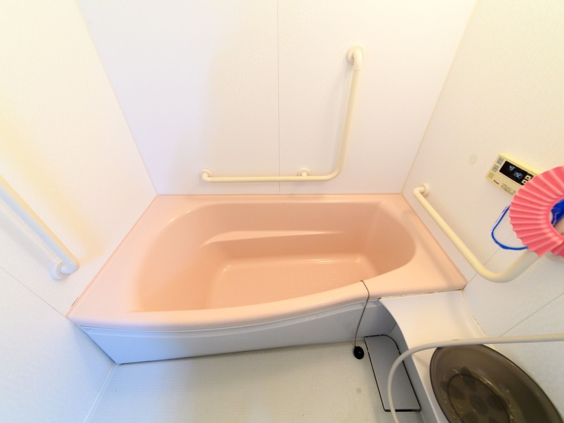白色の壁の浴室で、ピンクと白色のツートンカラーの浴槽である。壁には手すりや給湯器のリモコンが設置されている。