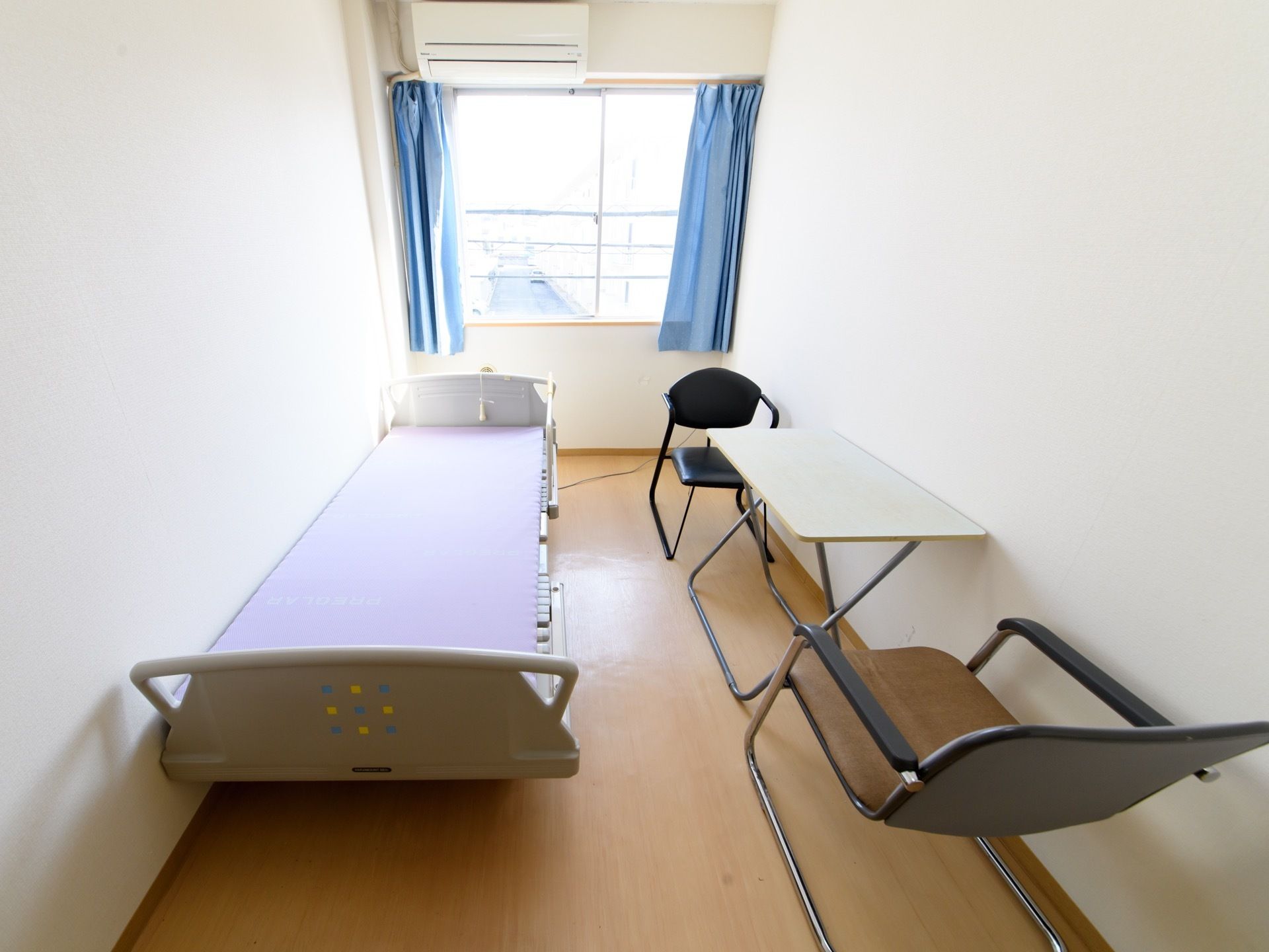 居室にベッドとテーブル、椅子が２脚置かれている。腰高窓にカーテンがかかり、窓の上の壁にエアコンが設置されている。
