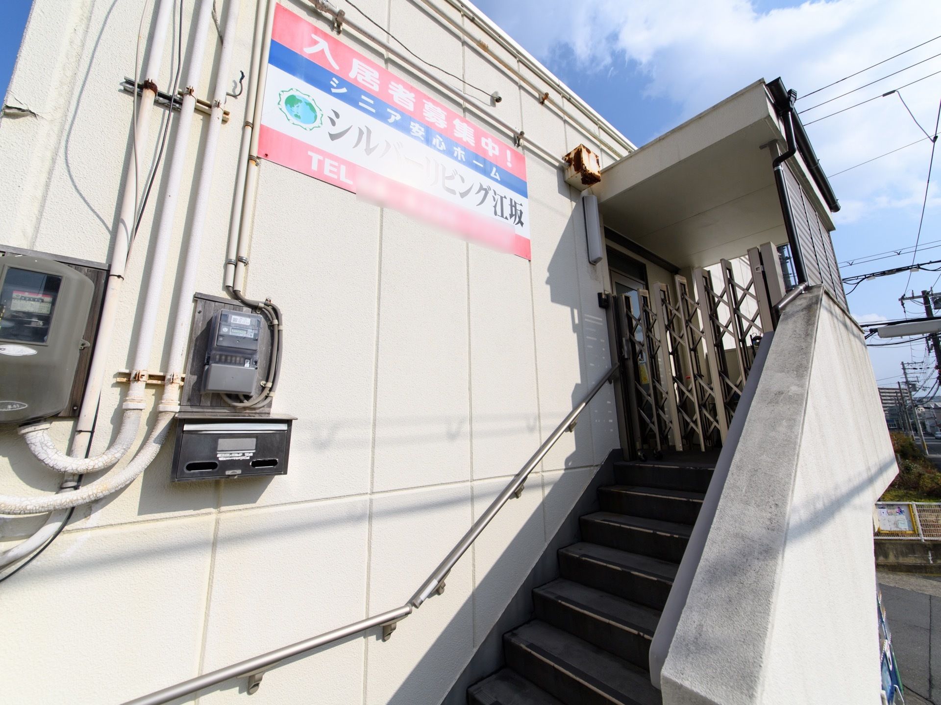 外から施設の二階部分である。手すりのついた階段がある。途中の壁に郵便受けが付いている。施設の看板も設置されている。