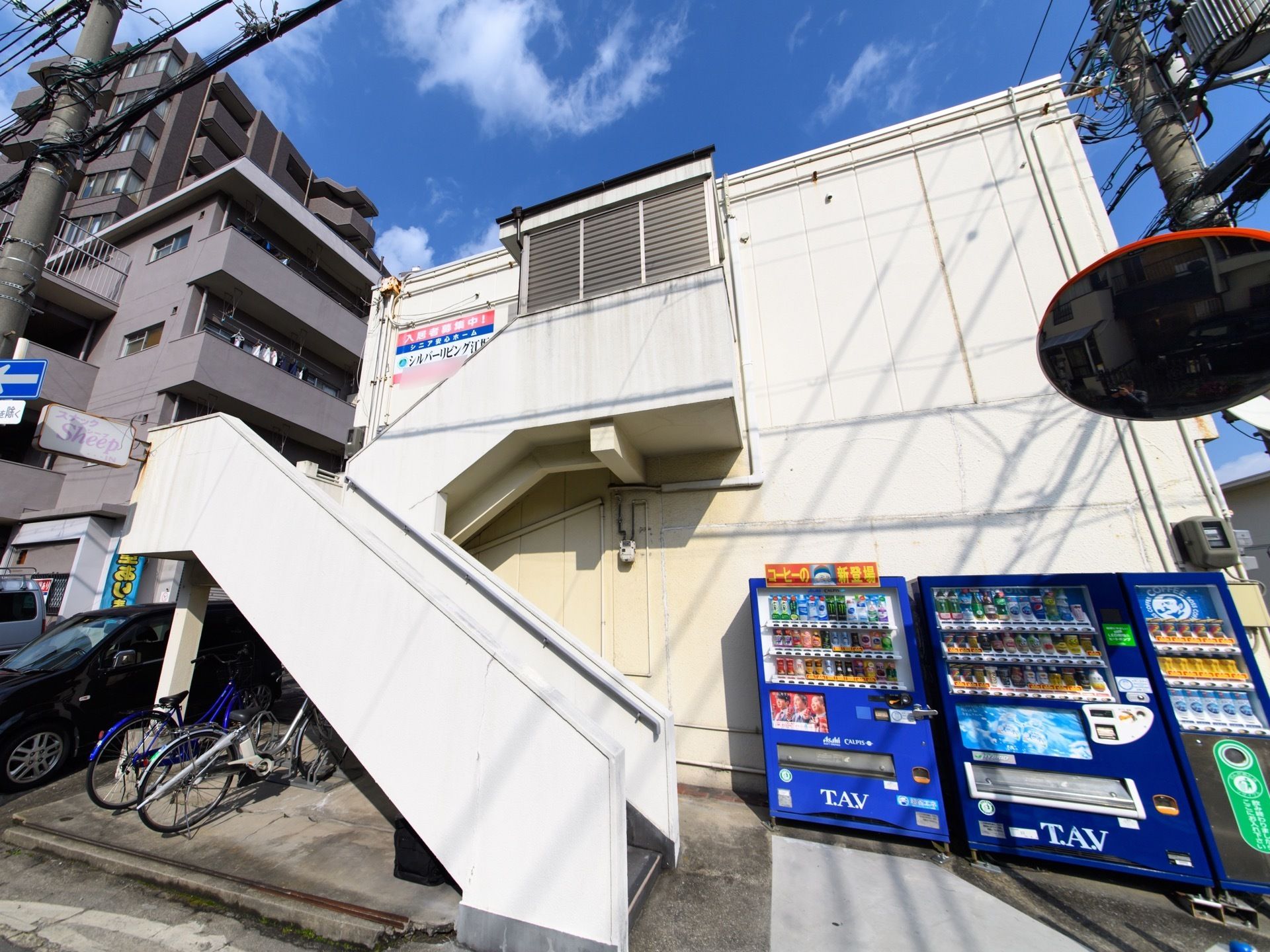 二階建ての建物で、手前に階段がある。階段下は自転車置き場になっており、右側の壁沿いに自動販売機が立っている。