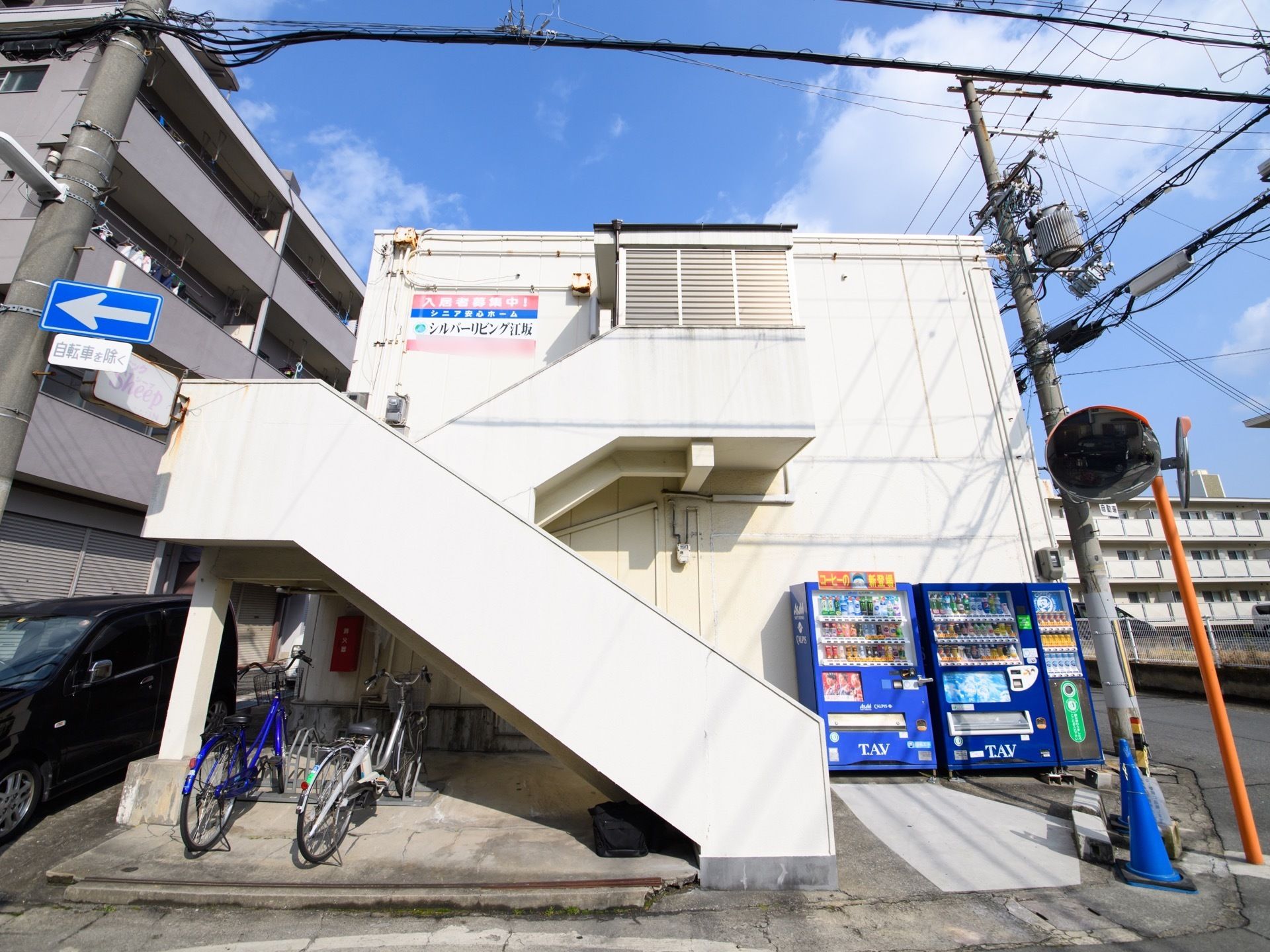 建物の手前に自動販売機が数台置かれている。壁面に施設の大きな看板が設置されている。二階への階段の下に自転車が停まっている。