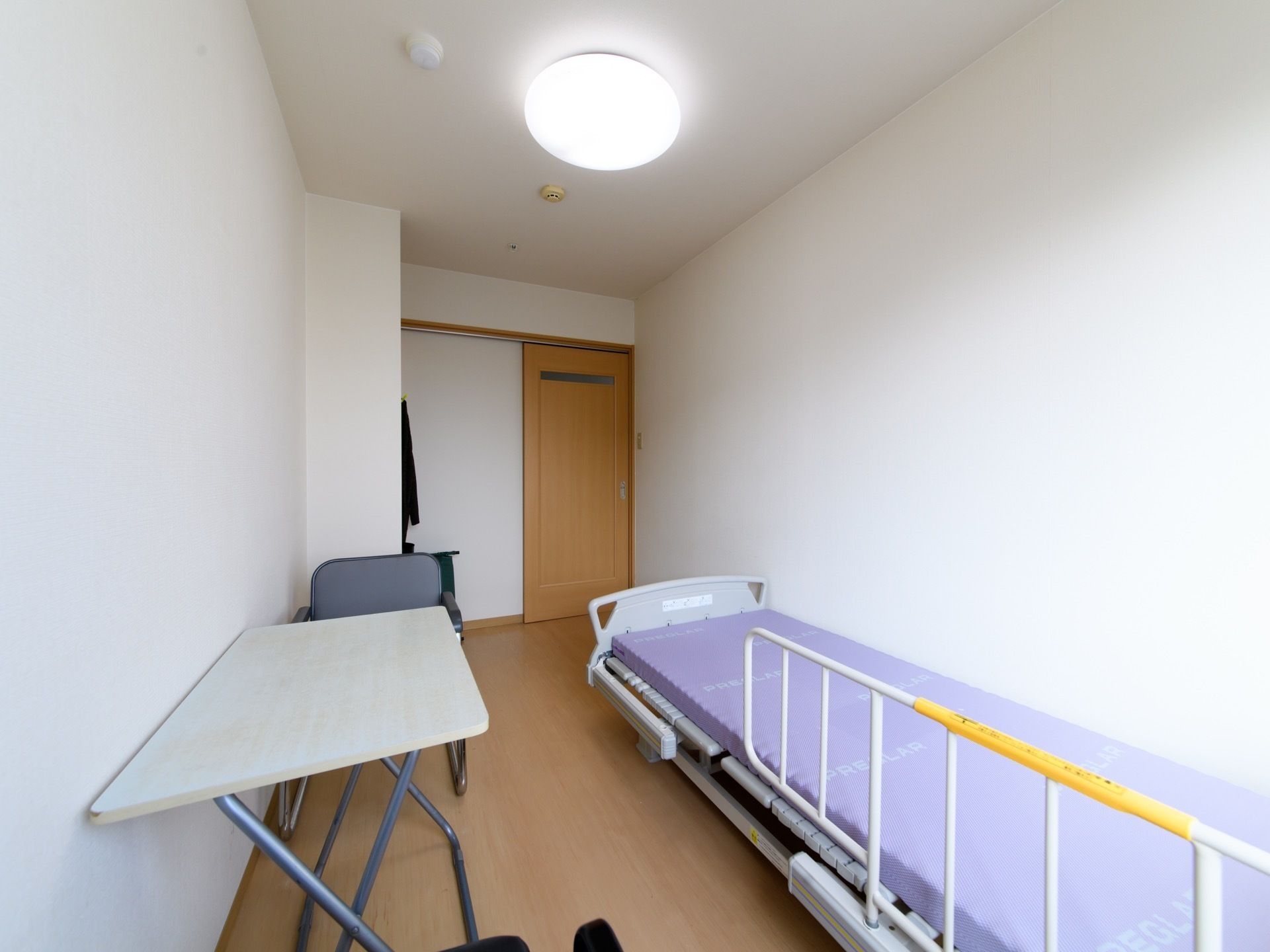 白い壁にフローリングの床の居室である。出入り口は引き戸で、ベッドや折りたたみ式テーブル、椅子が置かれている。