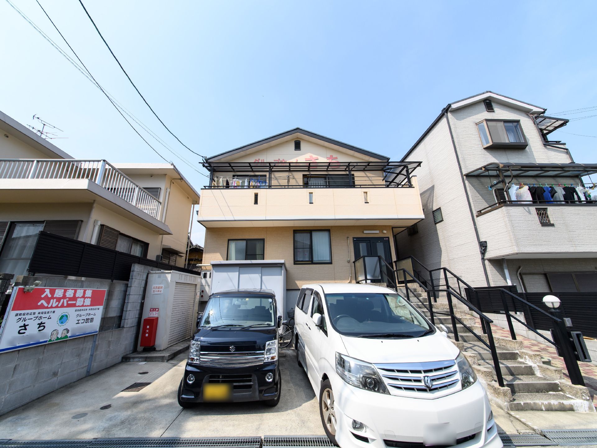 玄関のドアに続く階段があり、両側に黒色の手すりが備わっている。駐車場には入居者様募集やヘルパー募集の看板が掛けられている。