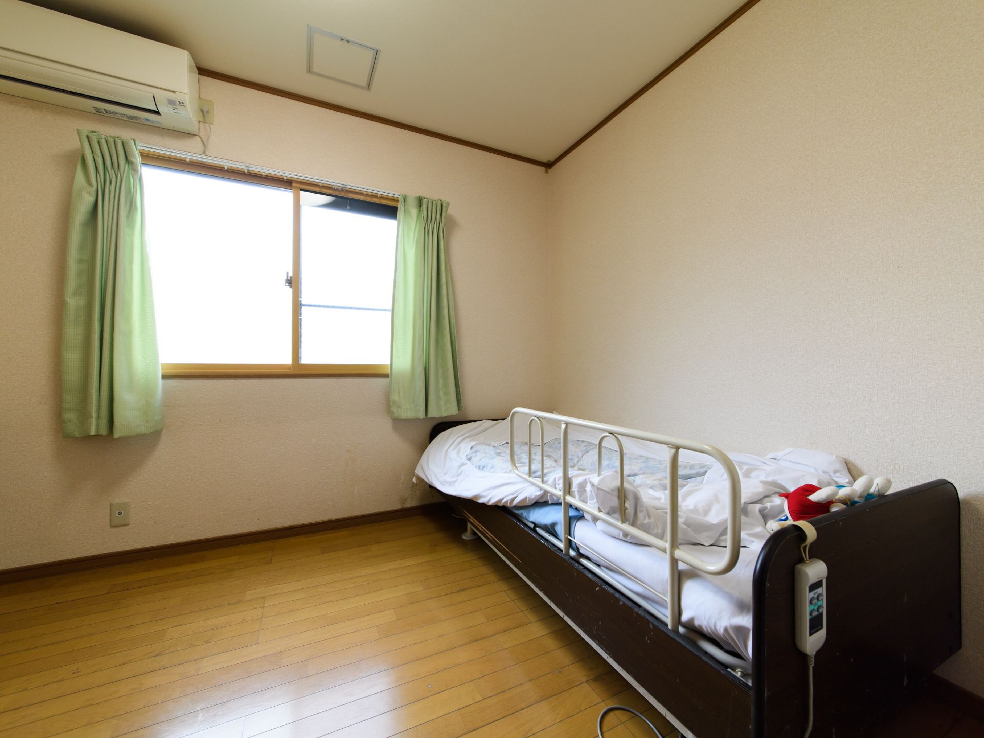 エアコンが完備されている。電動ベッドが配置されている。腰高窓があり、緑色のカーテンが取り付けてある。床はフローリングである。